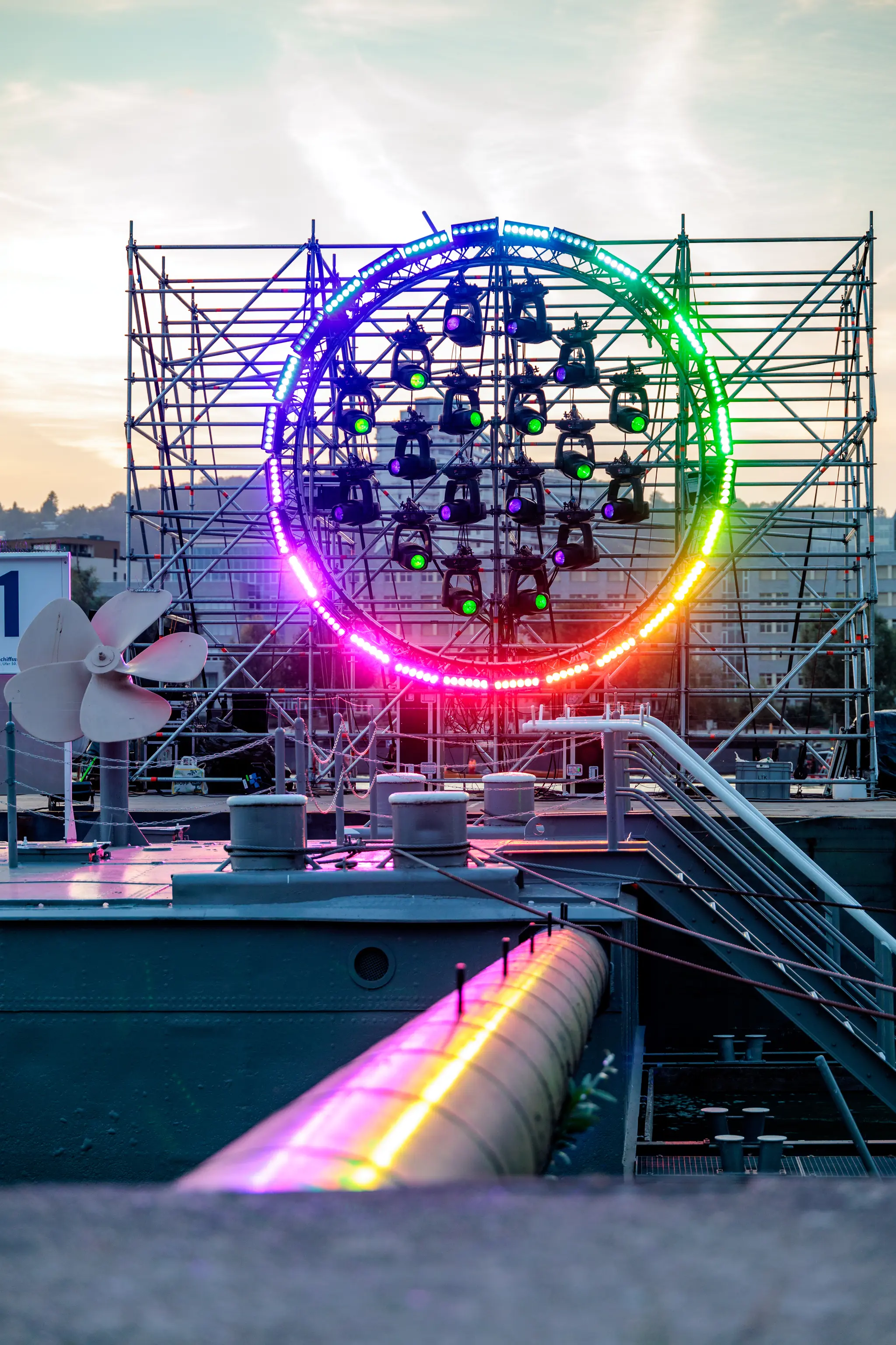 Farbenfroh leuchtende kreisförmige Lichtinstallation mit LEDs und Scheinwerfern auf einer Gerüstkonstruktion bei Dämmerung. Im Vordergrund sind Details eines Schiffes oder Docks wie ein Propeller, Rohre und Poller sichtbar