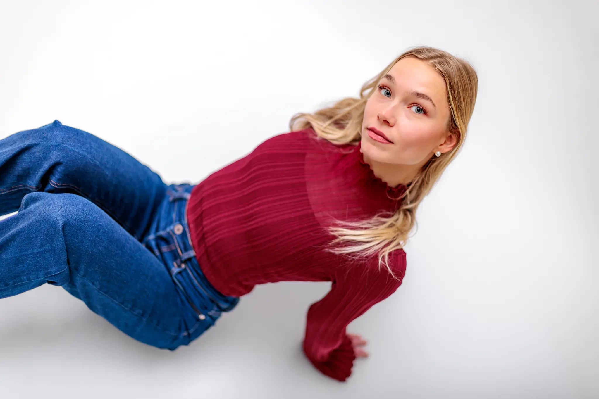 Ein modernes Studio-Portrait der blonden Kathrin, die in einem weinroten Rollkragenpullover und blauen Jeans für ein authentisches Lifestyle-Shooting auf weißem Hintergrund posiert