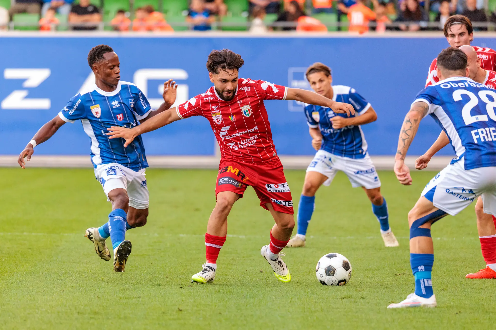Ein Welser Spieler treibt den Ball im Mittelfeld an und wird dabei von mehreren Linzer Spielern bedrängt; im Hintergrund leuchtet eine blaue Werbebande.