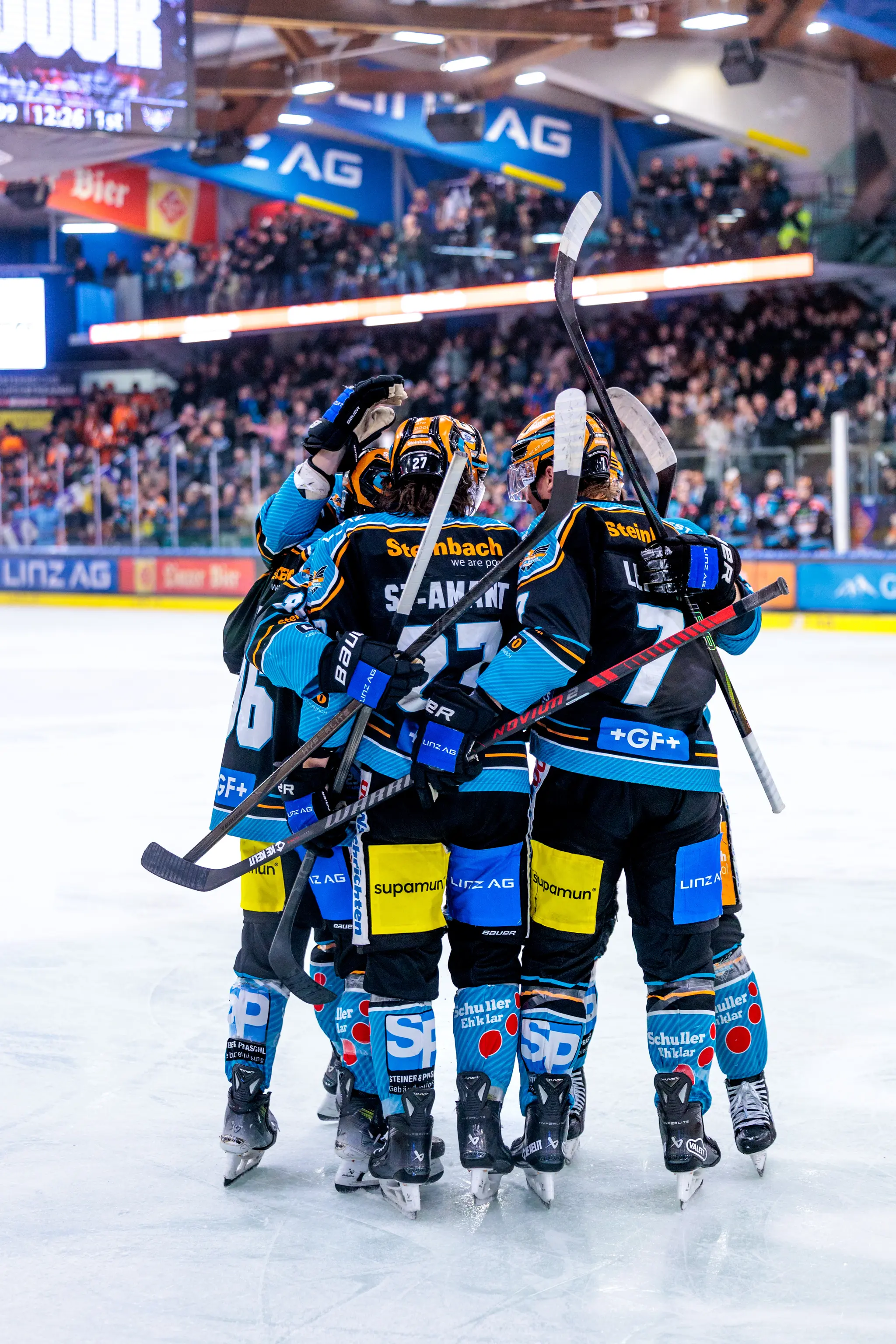 Eishockeyspieler der Black Wings Linz in ihren dunklen Heimtrikots feiern einen Torerfolg eng beieinander auf dem Eis 