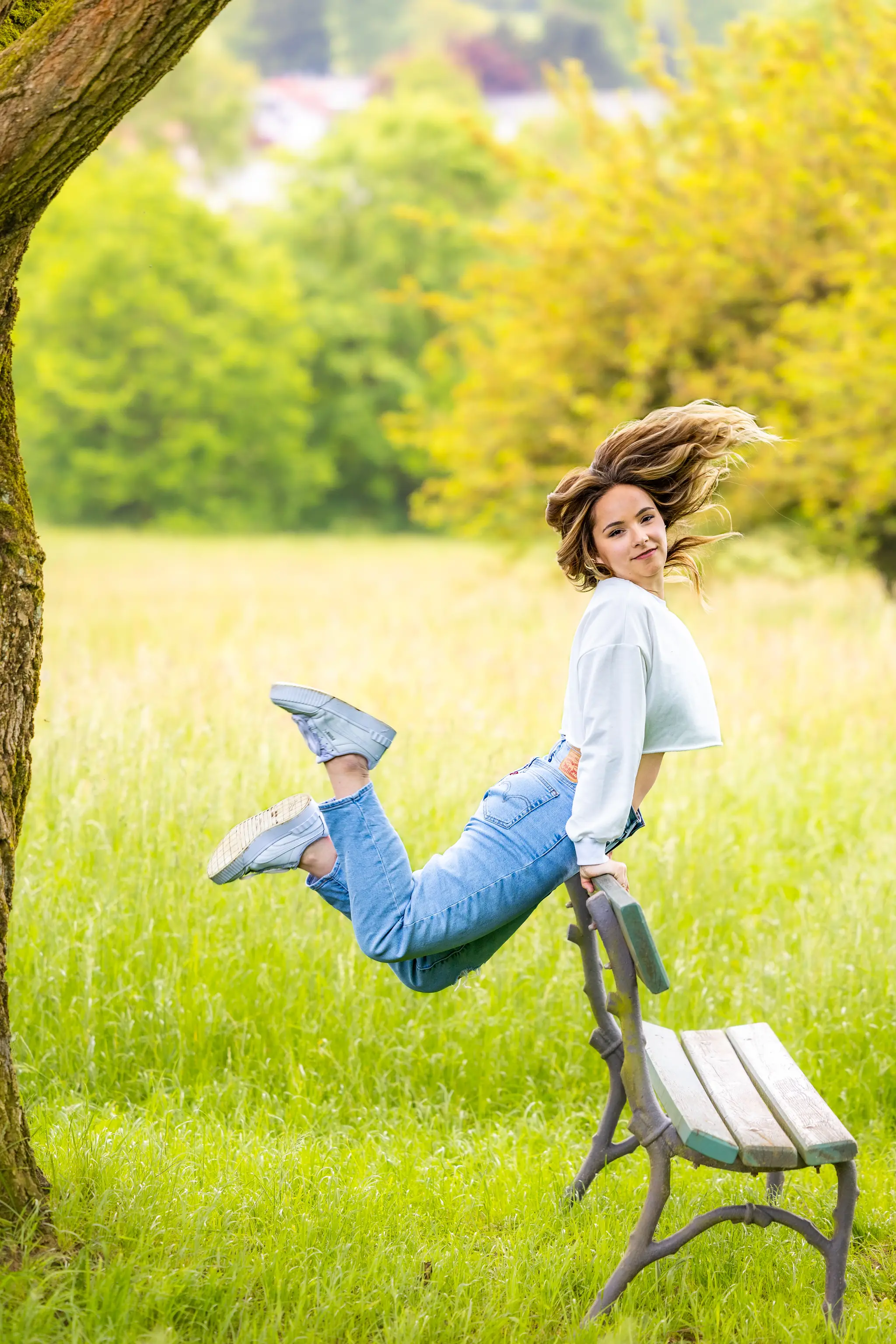 Eine junge Frau in Jeans und weißem Oberteil vollführt eine dynamische Pose auf einer Parkbank in einer grünen Parklandschaft, ihr Haar weht dynamisch