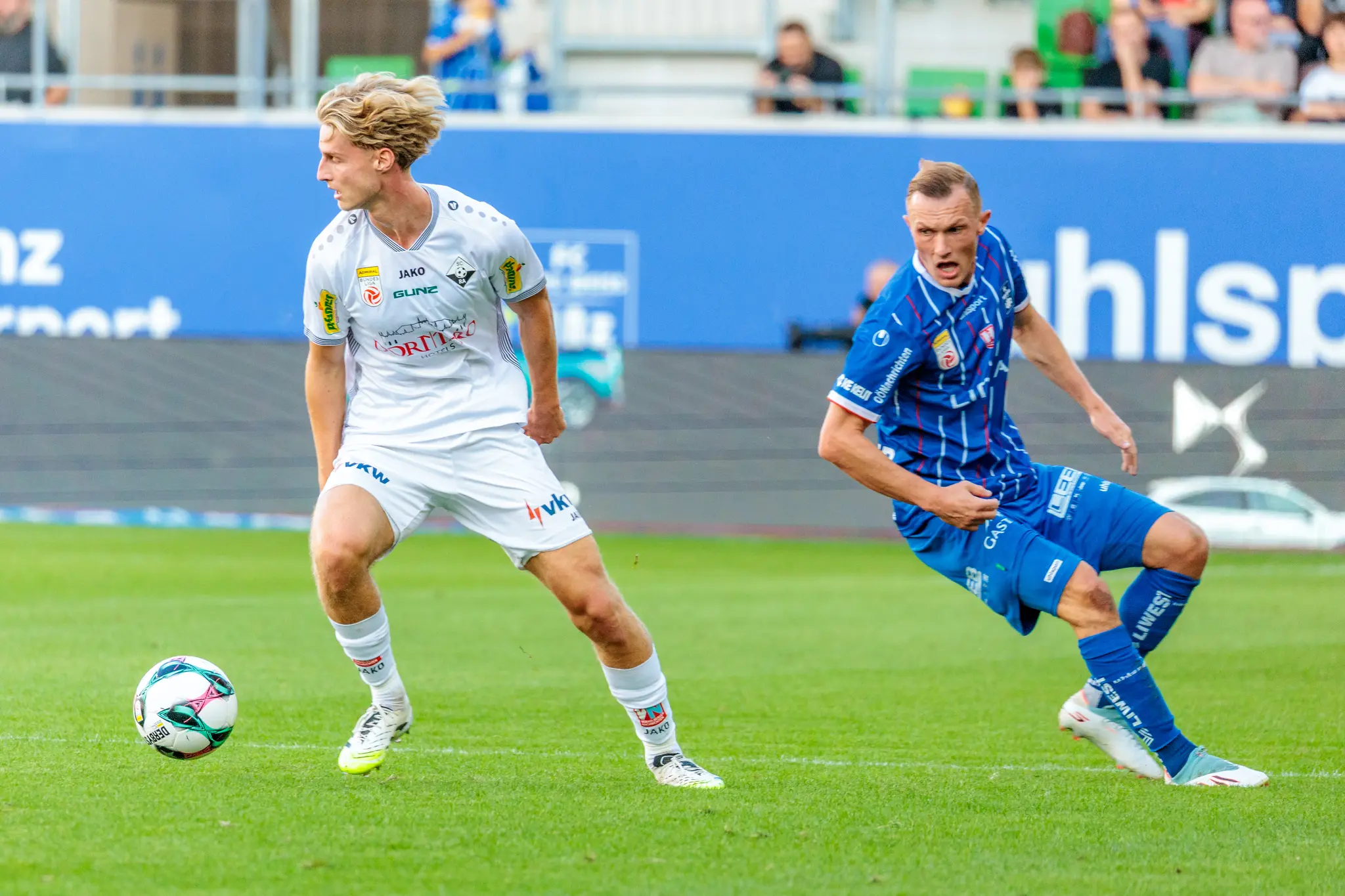 Ein Spieler des SCR Altach im weißen Trikot behauptet den Ball in einem dynamischen Zweikampf gegen einen heraneilenden Spieler von Blau-Weiß Linz