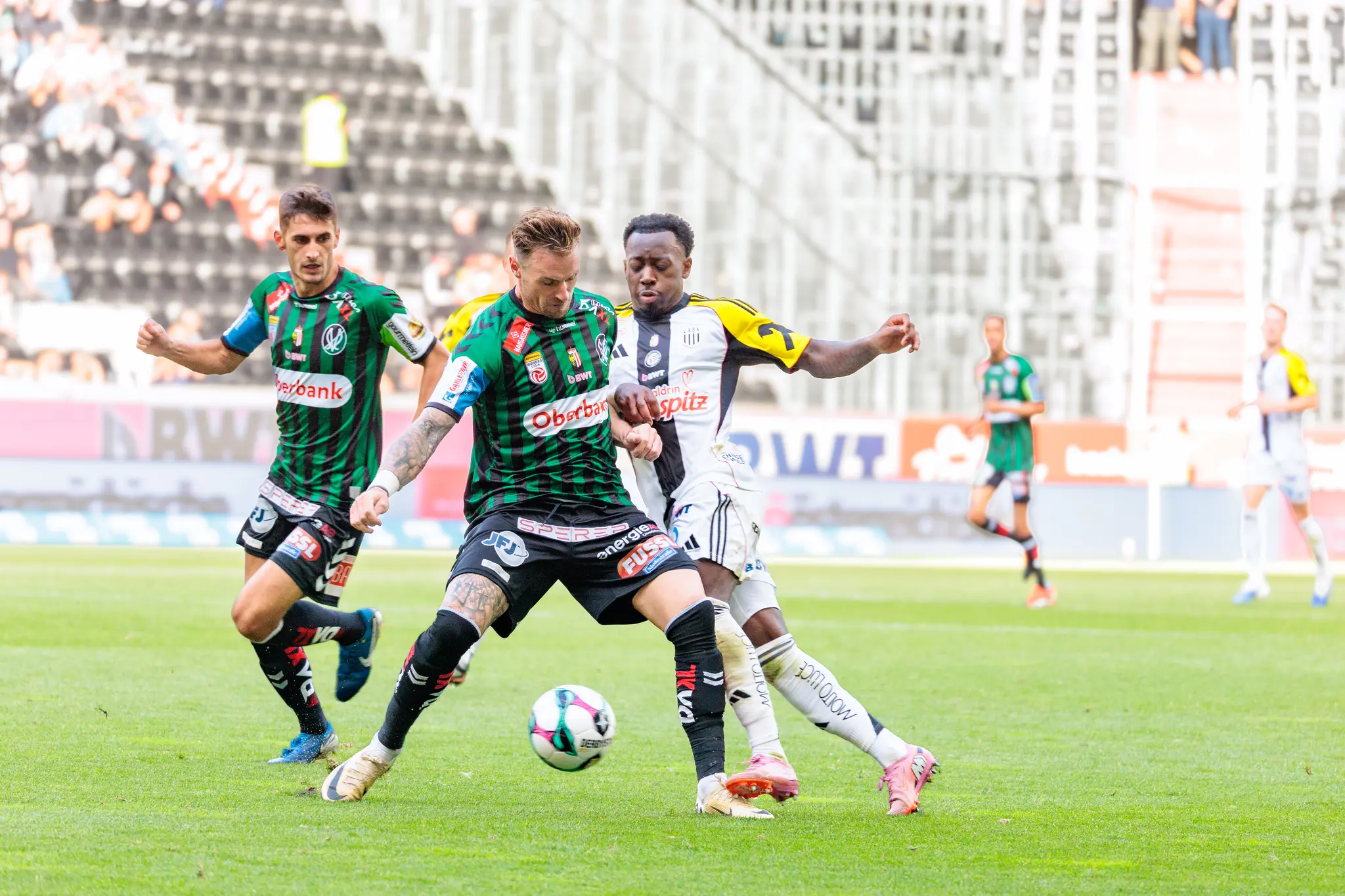 Ein Fußballspieler der SV Ried dribbelt den Ball im Mittelfeld, während ein Verteidiger des LASK ihn während eines Bundesligaspiels attackiert