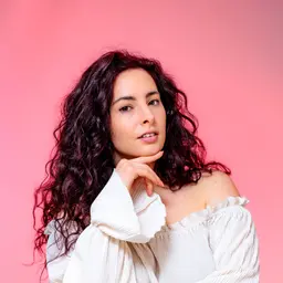 Porträt von Nicole Pecnik mit dunklem Lockenhaar und weißem Oberteil vor rosa Hintergrund