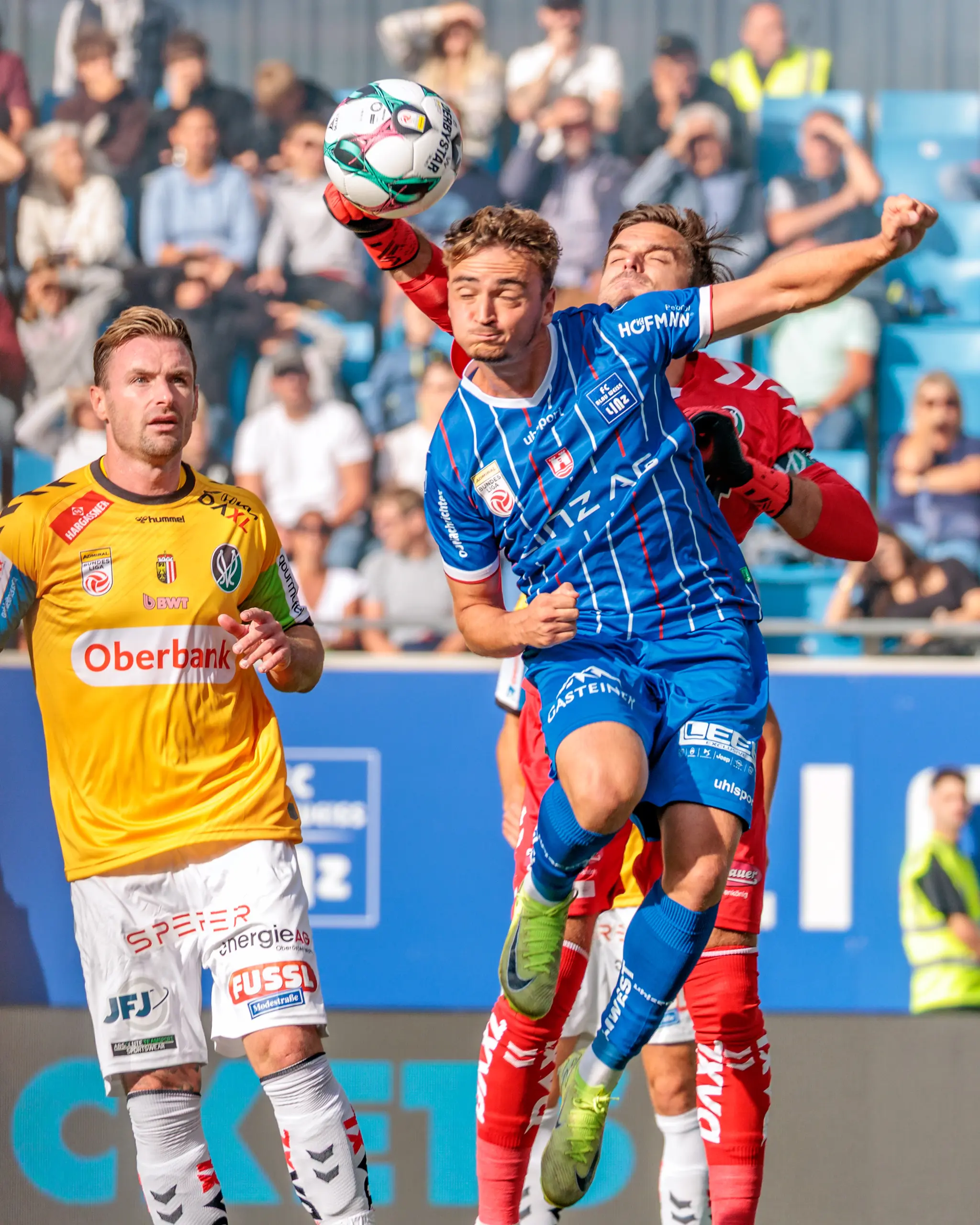 Der Torwart des SV Ried klärt den Ball mit einem Kopfball im Strafraum unter dem Druck eines Gegenspielers des Blau Weiß Linz, während weitere Spieler und Zuschauer im Hintergrund das Bundesliga-Derby verfolgen