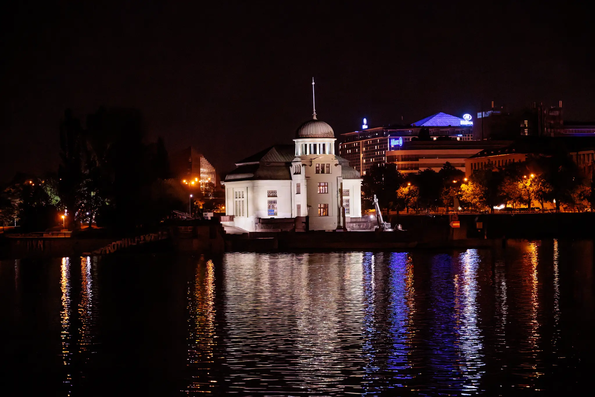 Das beleuchtete Wasserkraftwerk Stvanice mit Kuppel und Turm steht auf der Moldauinsel; die Lichter der Stadt spiegeln sich im Fluss.