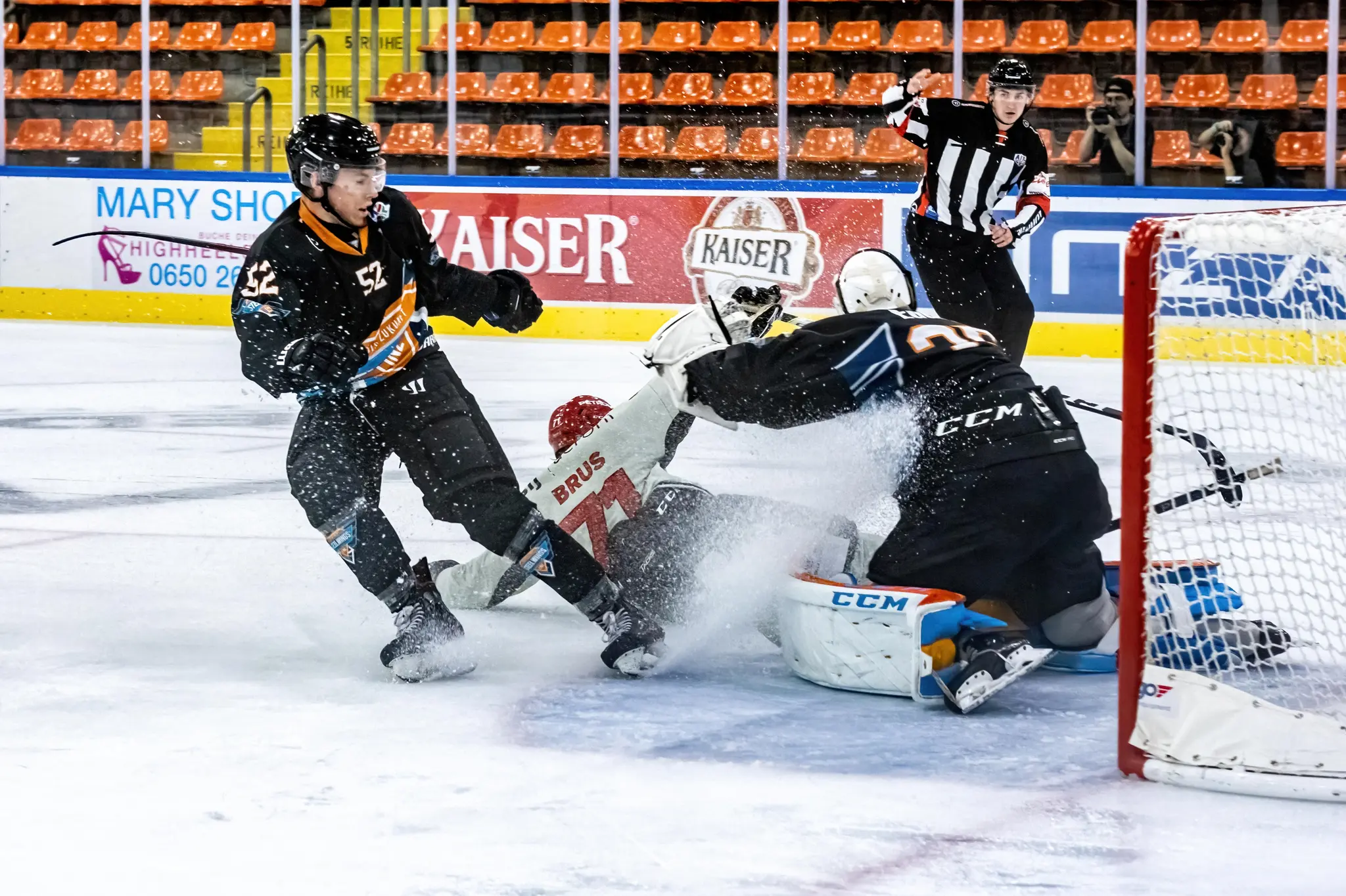 Ein Eishockeyspieler im dunklen Trikot ist in dynamischer Bewegung auf dem Eis vor dem Tor zu sehen, umringt von weiteren Spielern und dem Schiedsrichter