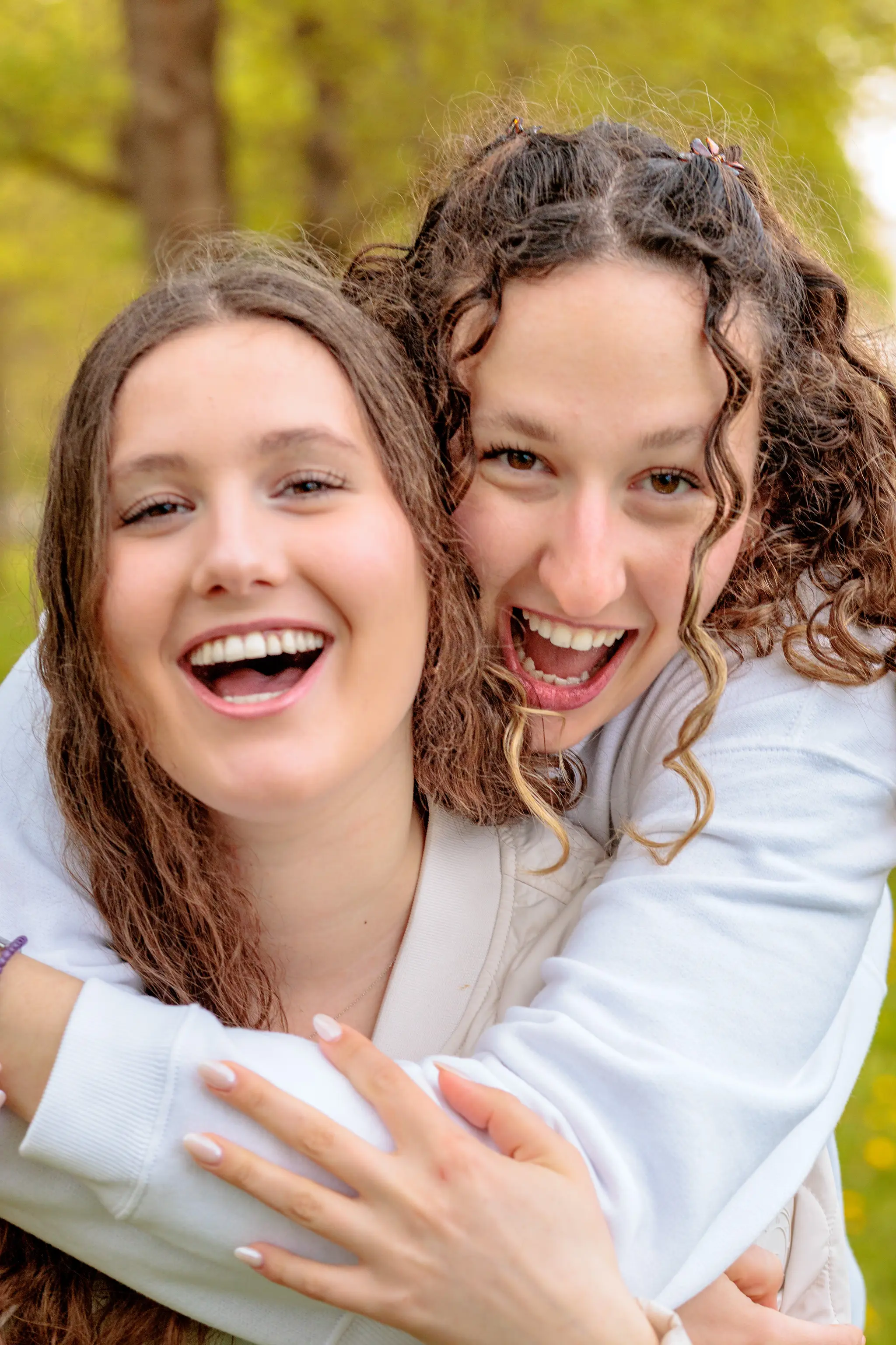 Zwei lachende Frauen mit braunem Haar umarmen sich liebevoll in einer hellen Frühlingslandschaft und strahlen Freude aus 