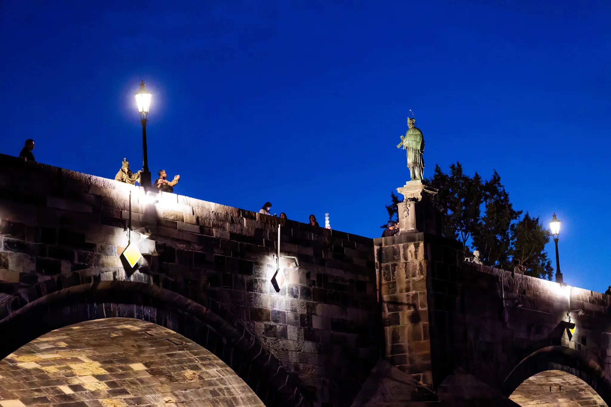 Auf der Karlsbrücke stehen Besucher zwischen den Statuen, einer fotografiert mit dem Handy; unter dem Brückenbogen fließt die Moldau in die Nacht.
