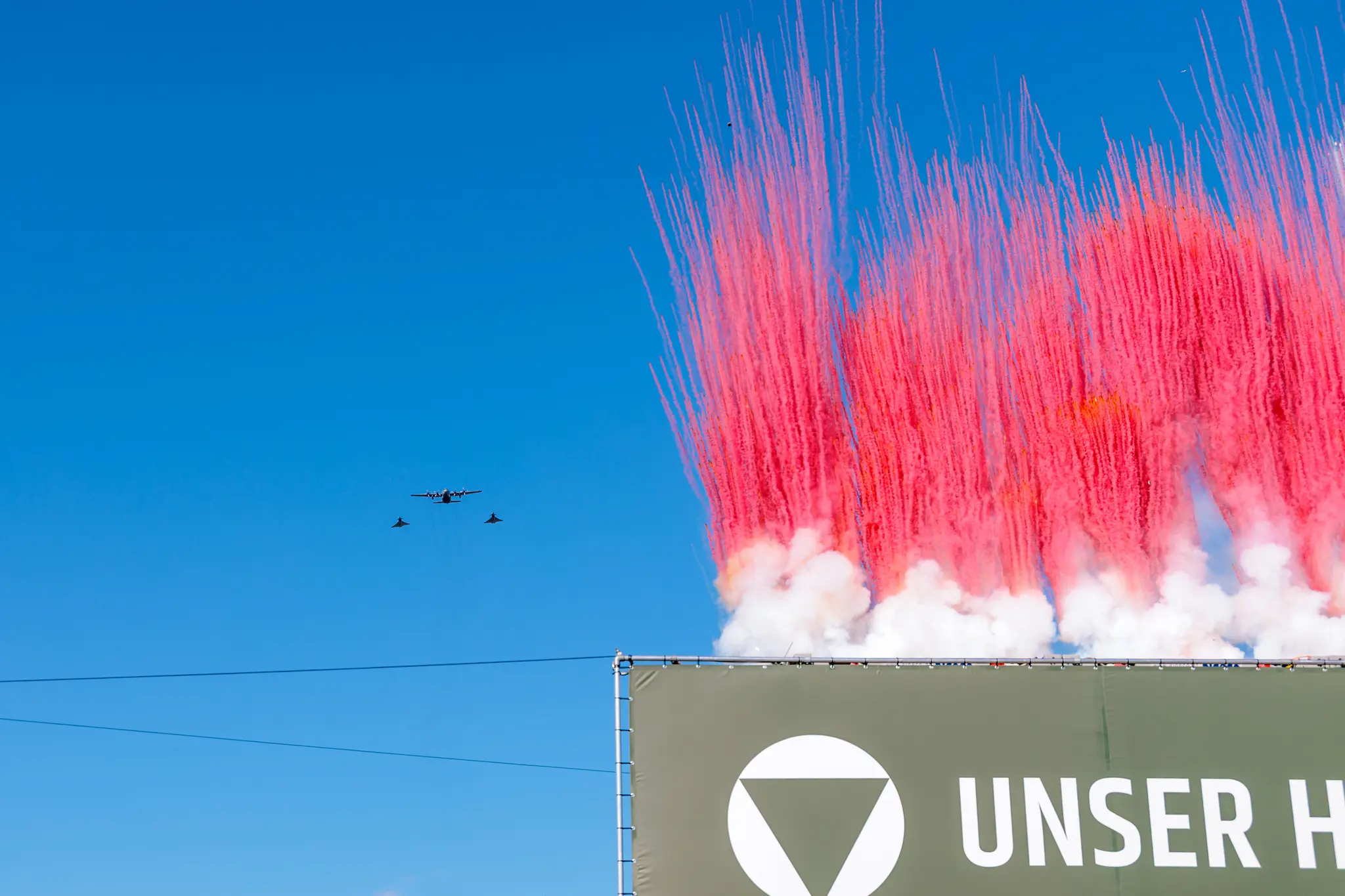 Eine Hercules-Transportmaschine und zwei Eurofighter fliegen über den Heldenplatz, während vor dem Banner „UNSER HEER“ dichte rot-weiße Rauchsäulen aufsteigen.
