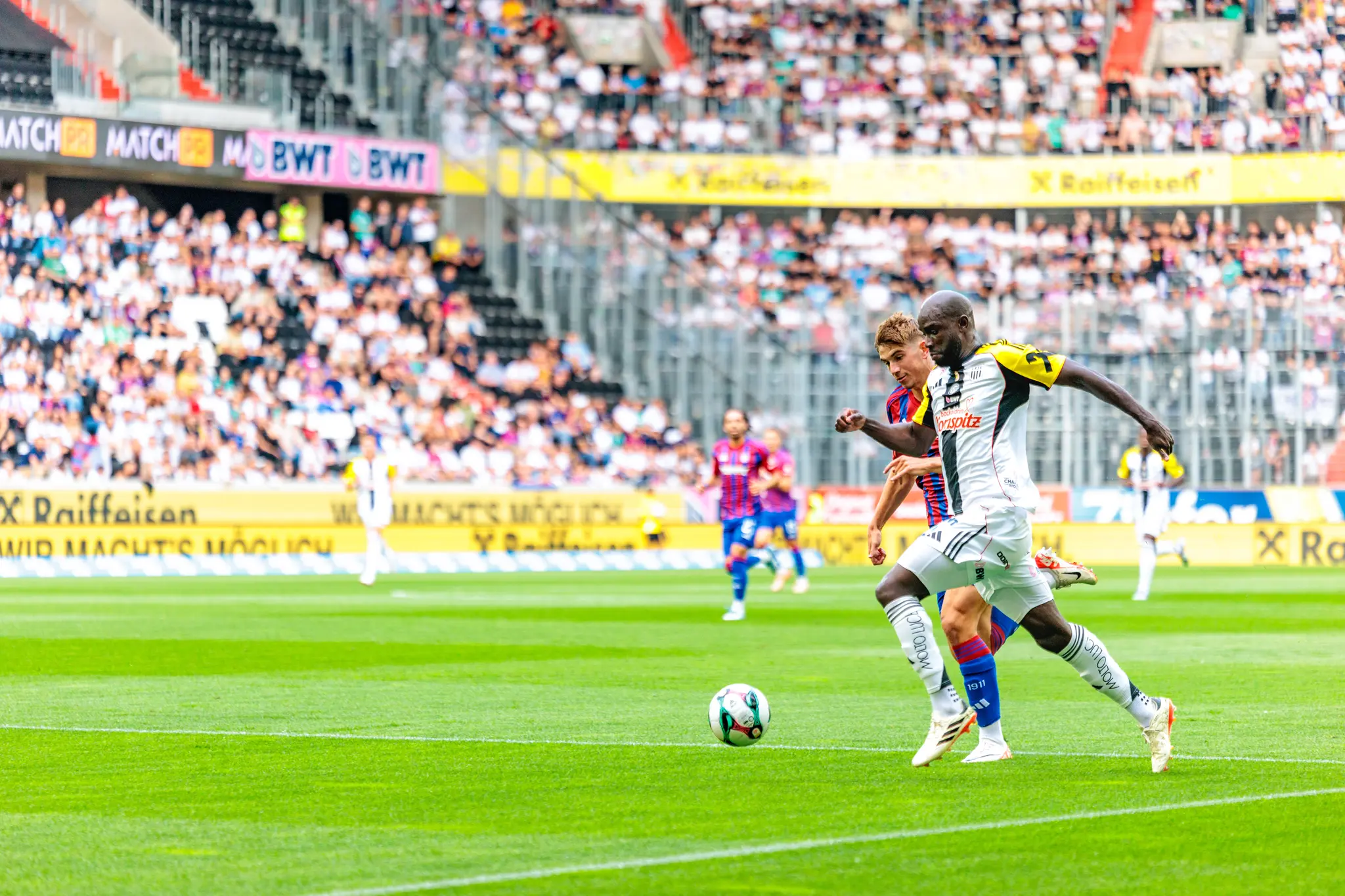 Zwei Fußballspieler im Duell um den Ball während eines Spiels in der Raiffeisen Arena, umgeben von Fans 