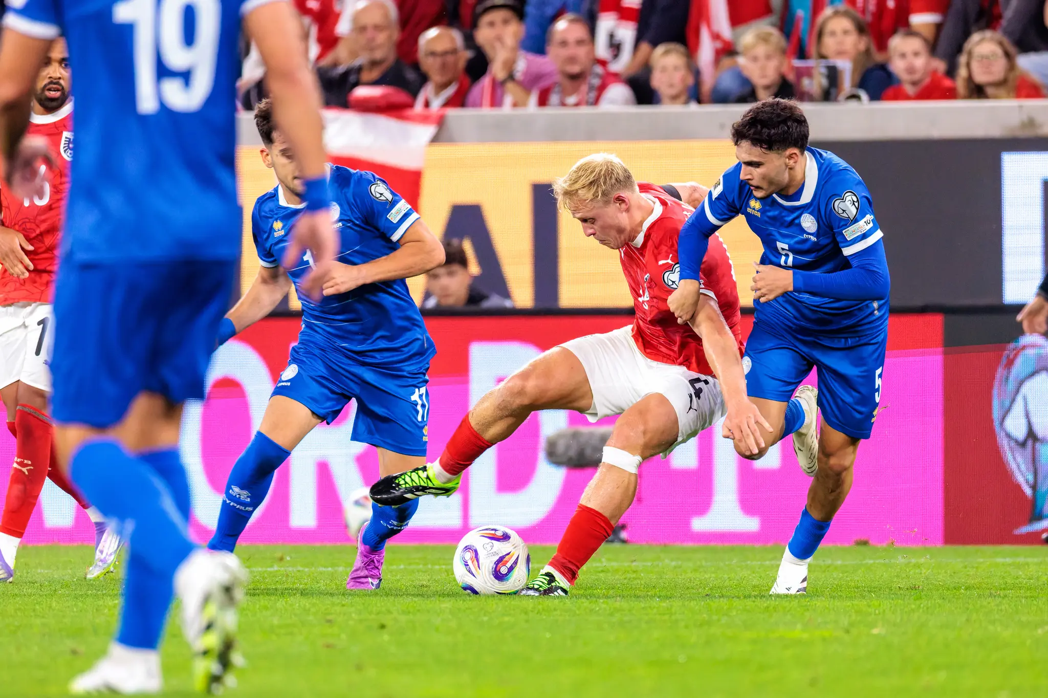 Xaver Schlager von Österreich im roten Trikot ringt im Zweikampf mit Charalampos Charalampous von Zypern im blauen Trikot um den Ball, während Konstantinos Laifis im Vordergrund das geschehen beobachtet