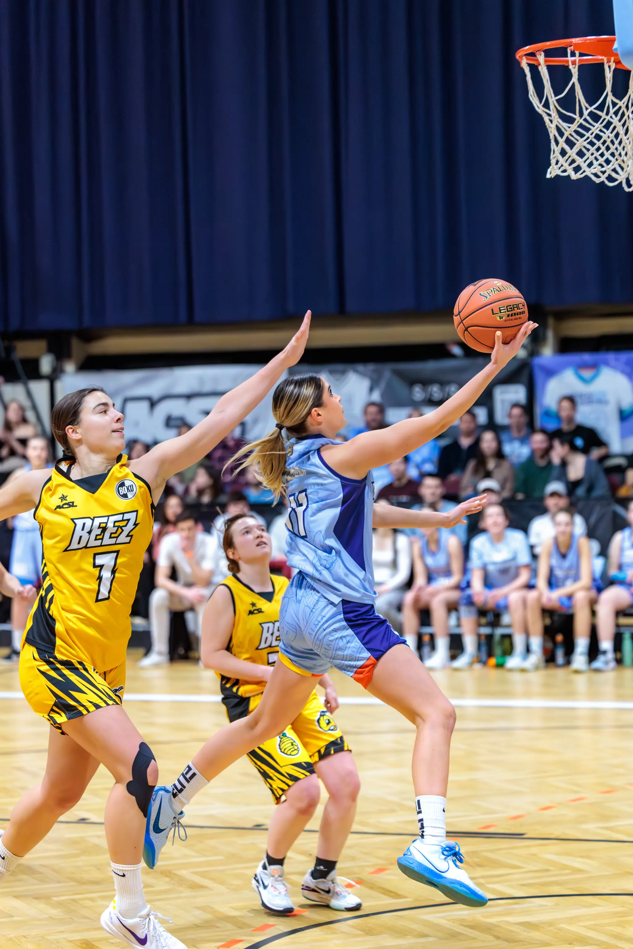 Frauen-Basketballspiel: Eine Spielerin in hellblauem Trikot wirft den Ball auf den Korb, während eine Gegnerin in gelbem Trikot mit der Nummer 7 verteidigt