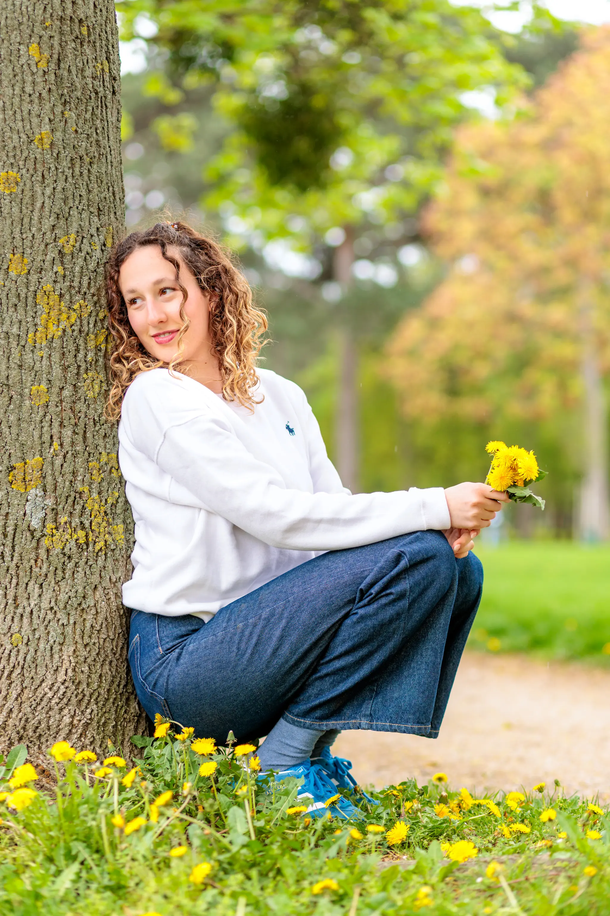 Eine lächelnde junge Frau mit lockigem Haar hockt im Frühlingspark an einem Baumstamm, hält Löwenzahn und trägt ein weißes Sweatshirt mit blauer Logostickerei und Jeans 