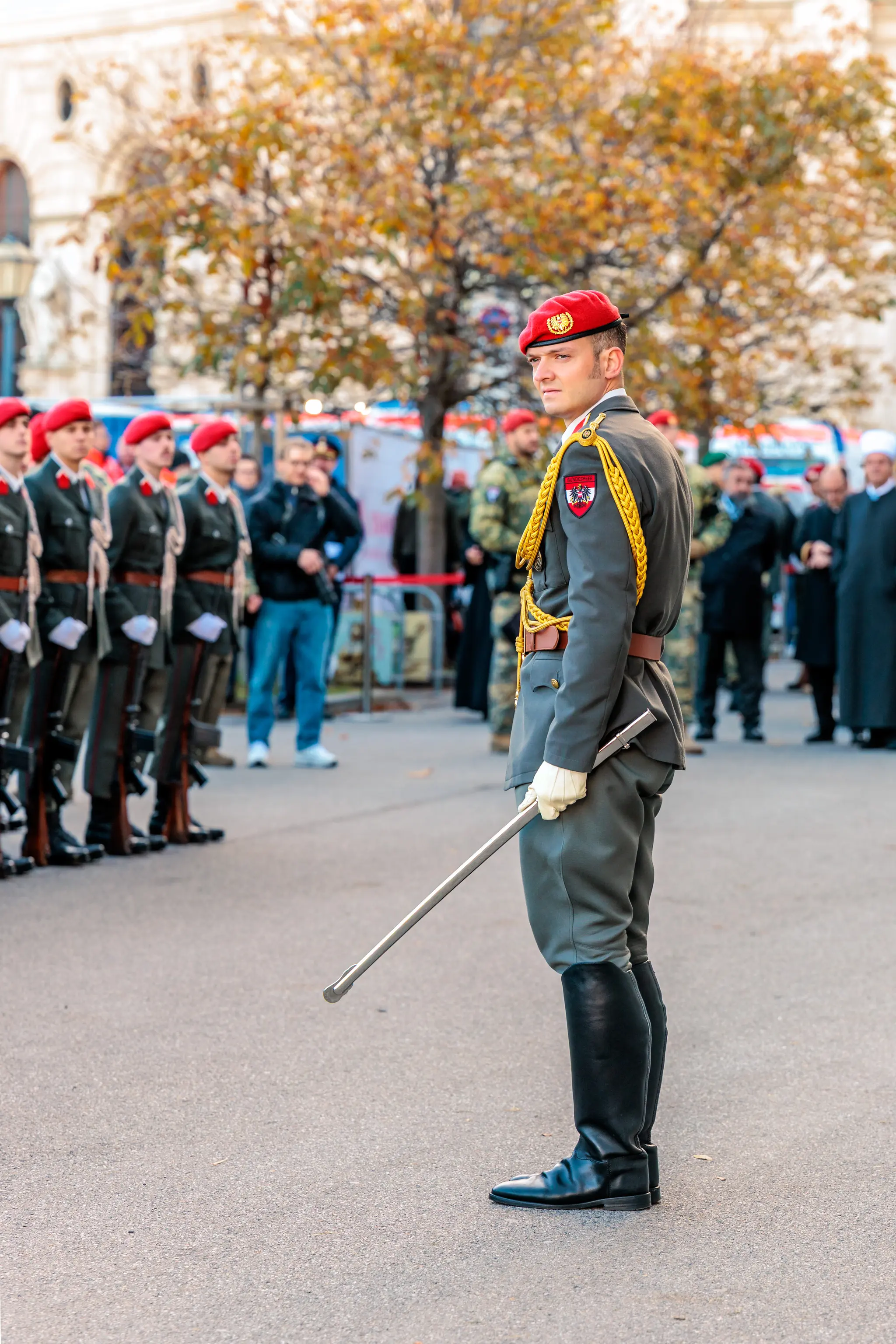 Ein Offizier der Garde steht im Profil mit gezogenem Säbel auf dem Heldenplatz, im Hintergrund wartet eine Formation von Soldaten mit roten Baretten.