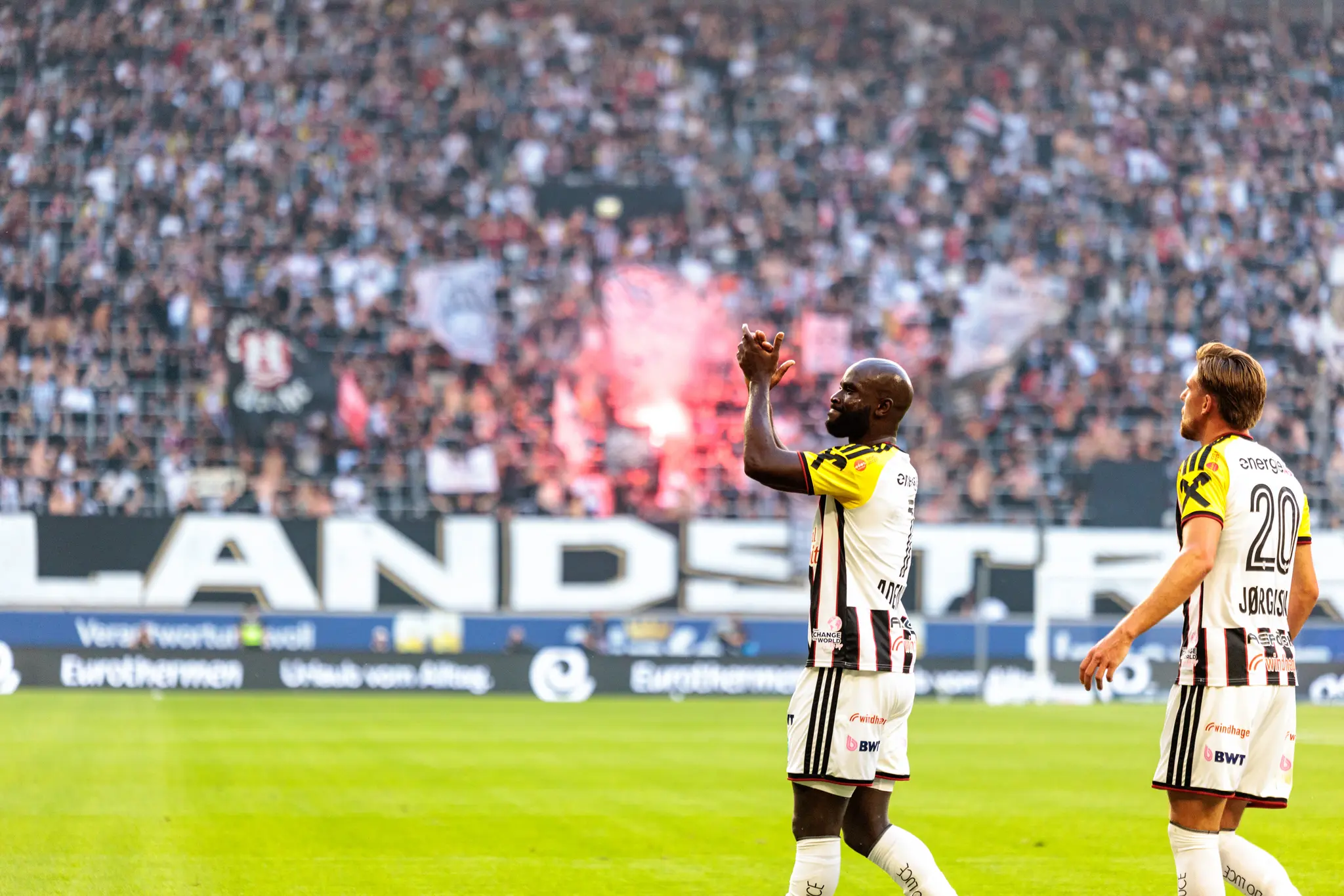 Adeniran applaudiert in Richtung Tribüne, im Hintergrund steigen roter Rauch und Pyrotechnik der Fans auf