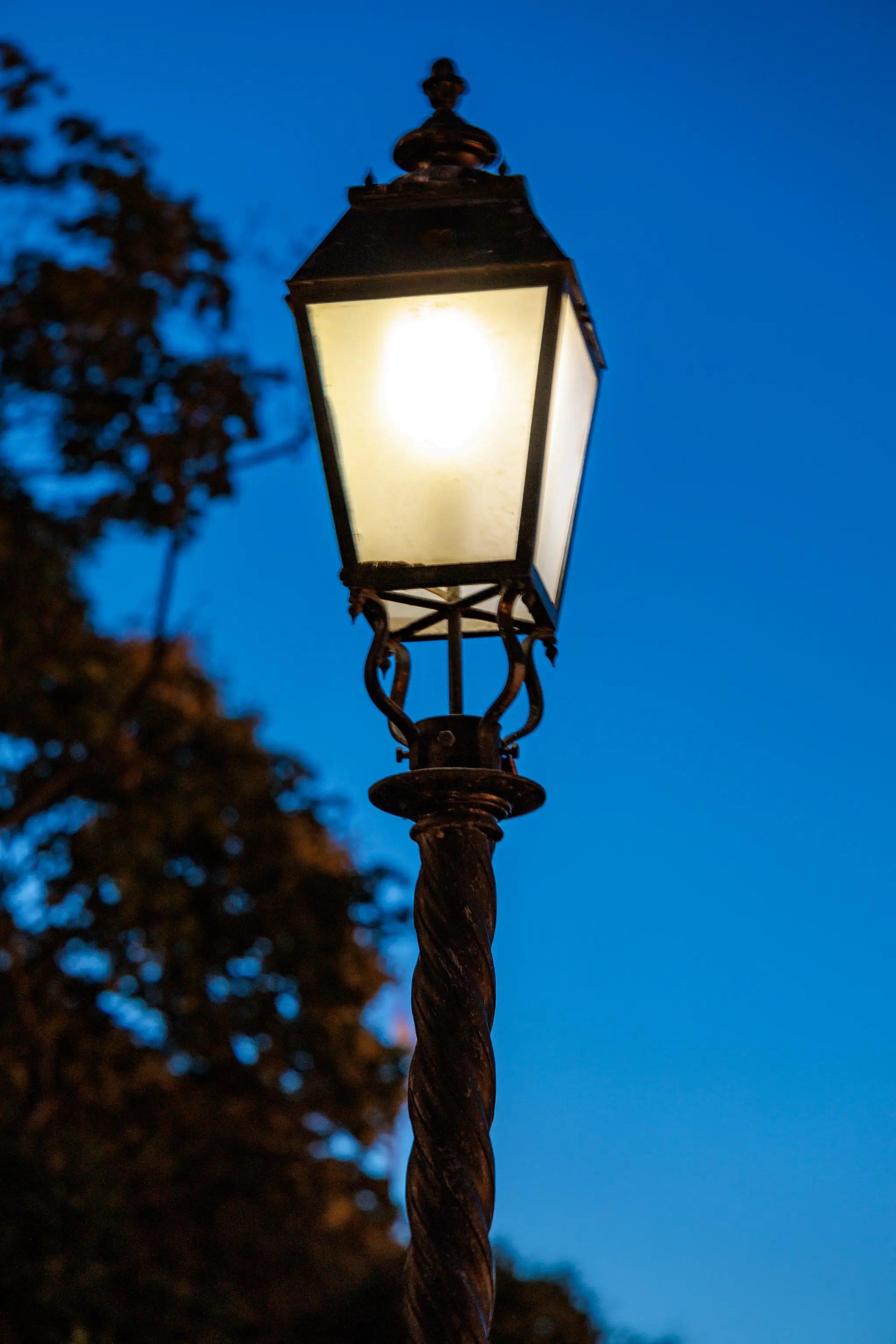 Eine klassische, beleuchtete Straßenlaterne mit gedrehtem Mast vor einem tiefblauen Abendhimmel und unscharfen Bäumen. 