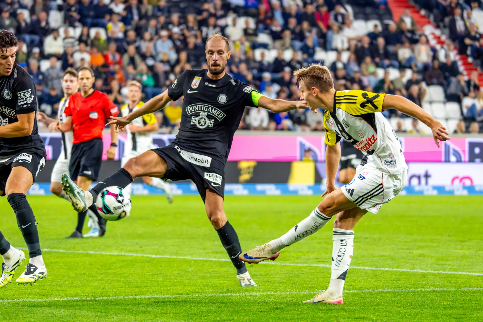 Ein Fußballspieler in dunklem Trikot schießt den Ball während eines Zweikampfs mit einem LASK-Spieler in hellem Trikot in einem Stadion