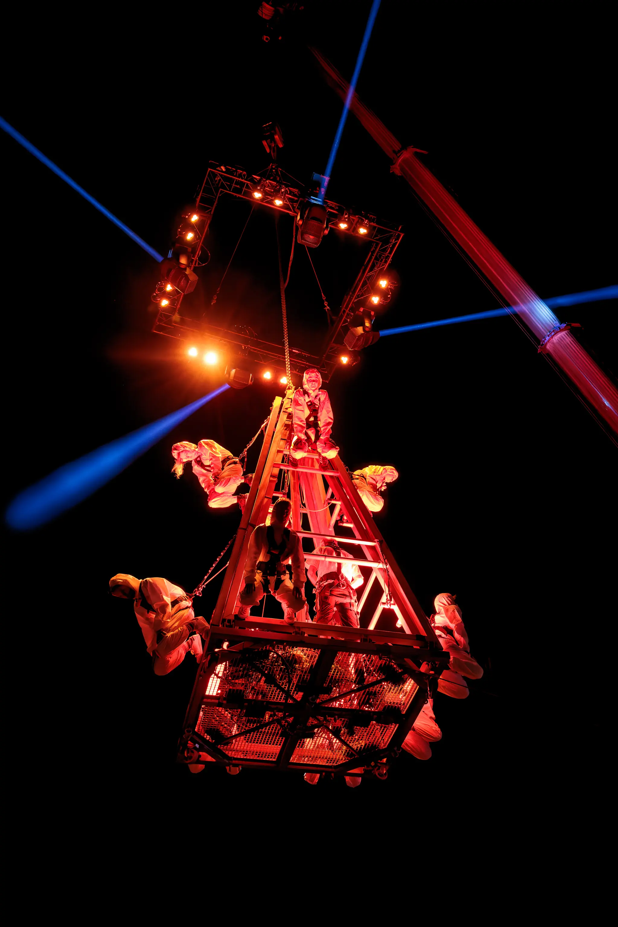 Mehrere Personen in Sicherheitsgurten auf einem schwebenden, rot beleuchteten Metallgerüst mit Leiterstruktur, umgeben von blauen Lichtstrahlen während der Linzer Klangwolke 2024. 