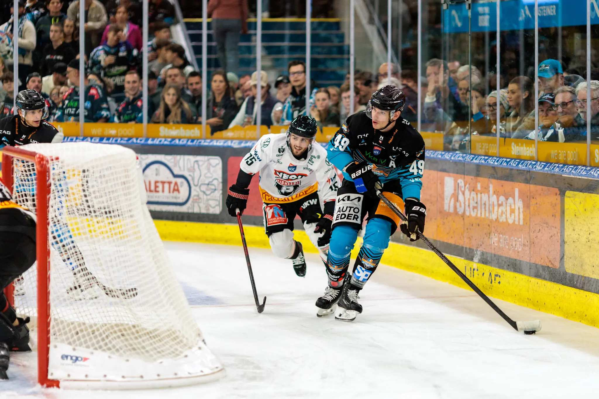 Eishockeyspieler Graham Knott von den Steinbach Black Wings Linz im teal-schwarzen Trikot mit der Nummer 49 führt den Puck über das Eis, während Mark Mussbacher der Pioneers Vorarlberg mit der Nummer 15 ihm folgt