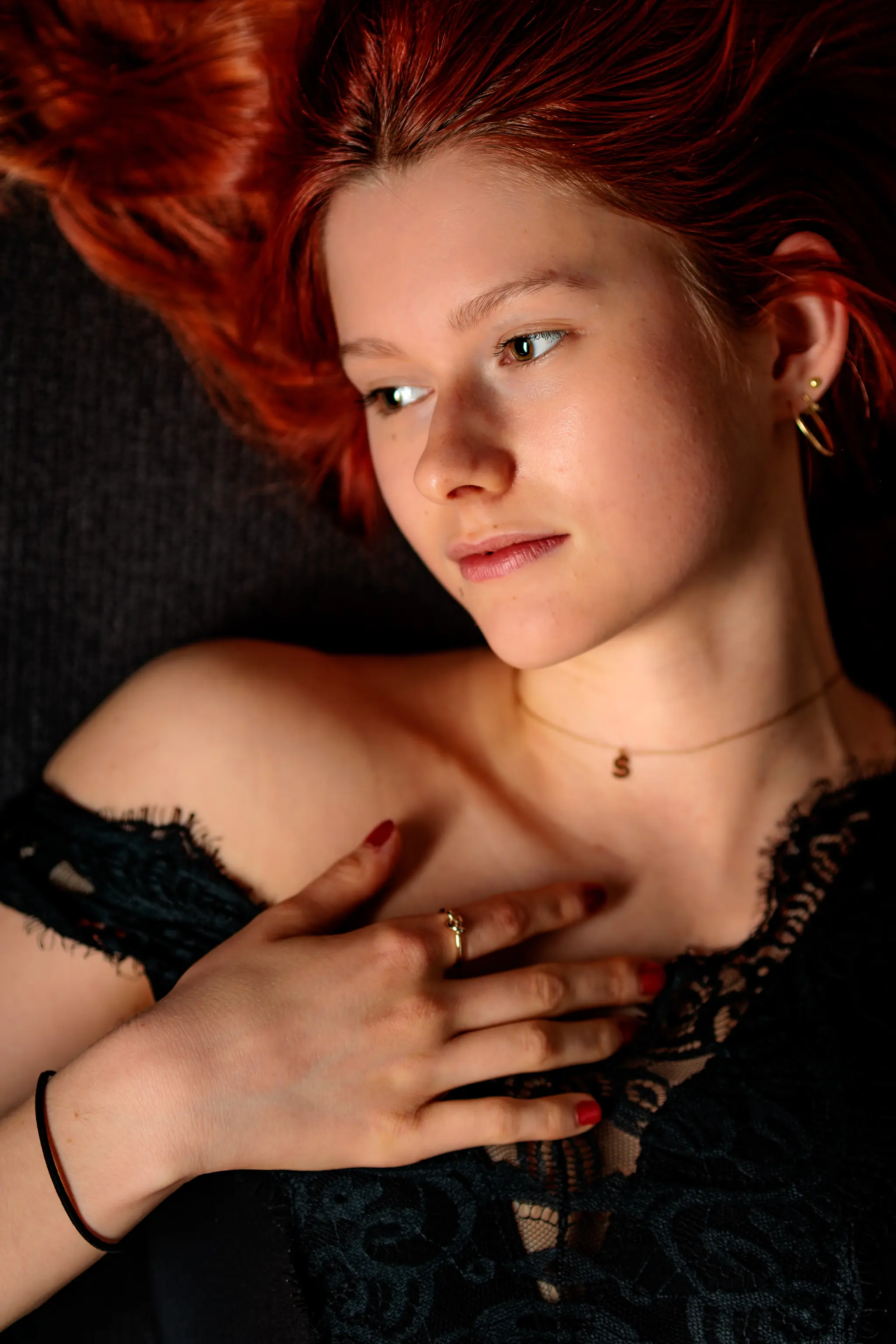 Verträumtes Portrait einer jungen rothaarigen Frau in schwarzer Spitze, die liegend sanft ihre Hand mit rotem Nagellack und goldenem Ring auf dem Dekolleté hält