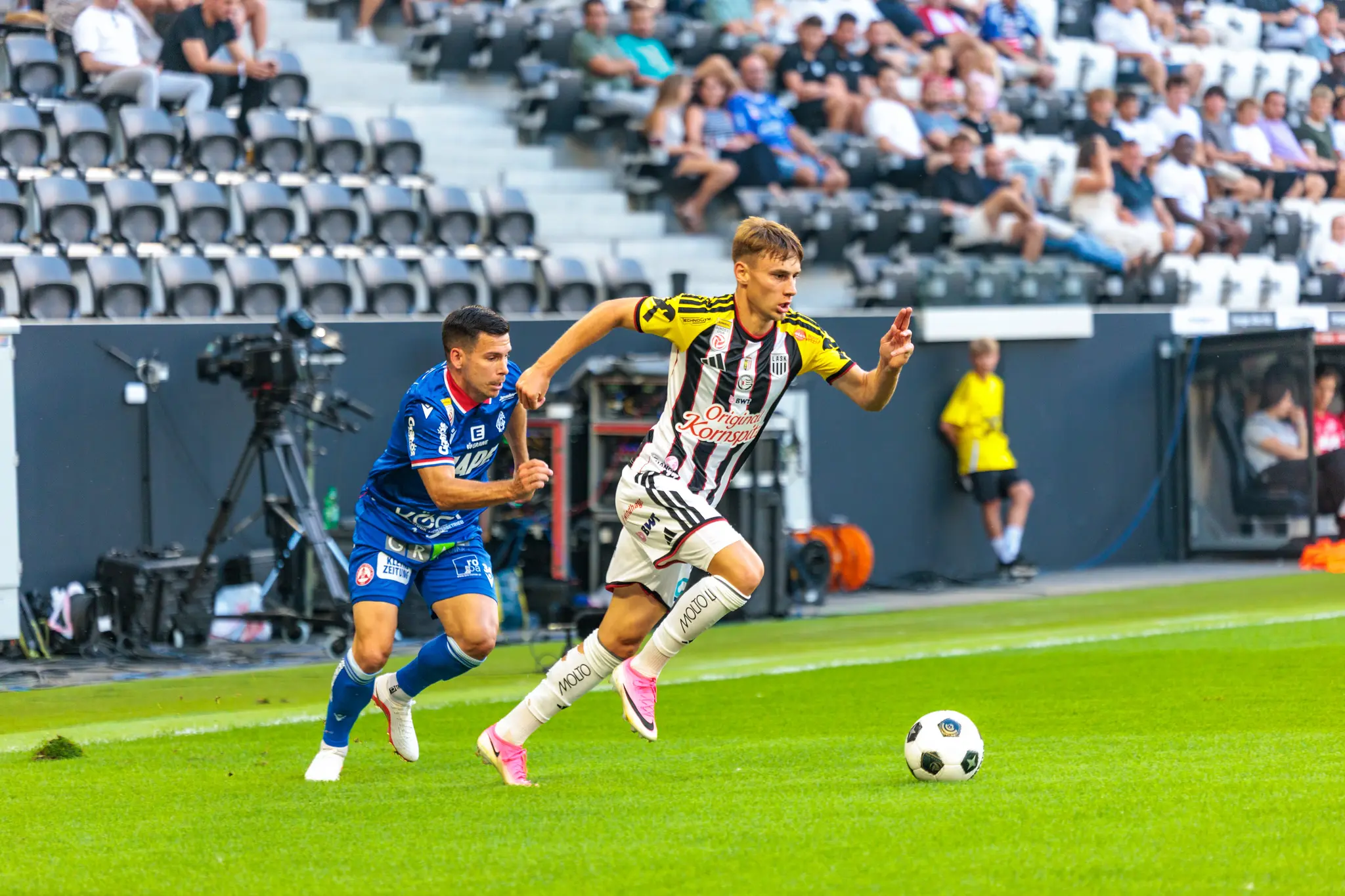 Junger LASK-Spieler dribbelt mit dem Ball an der linken Spielfeldseite, während ein GAK-Verteidiger im blauen Trikot hinter ihm herläuft
