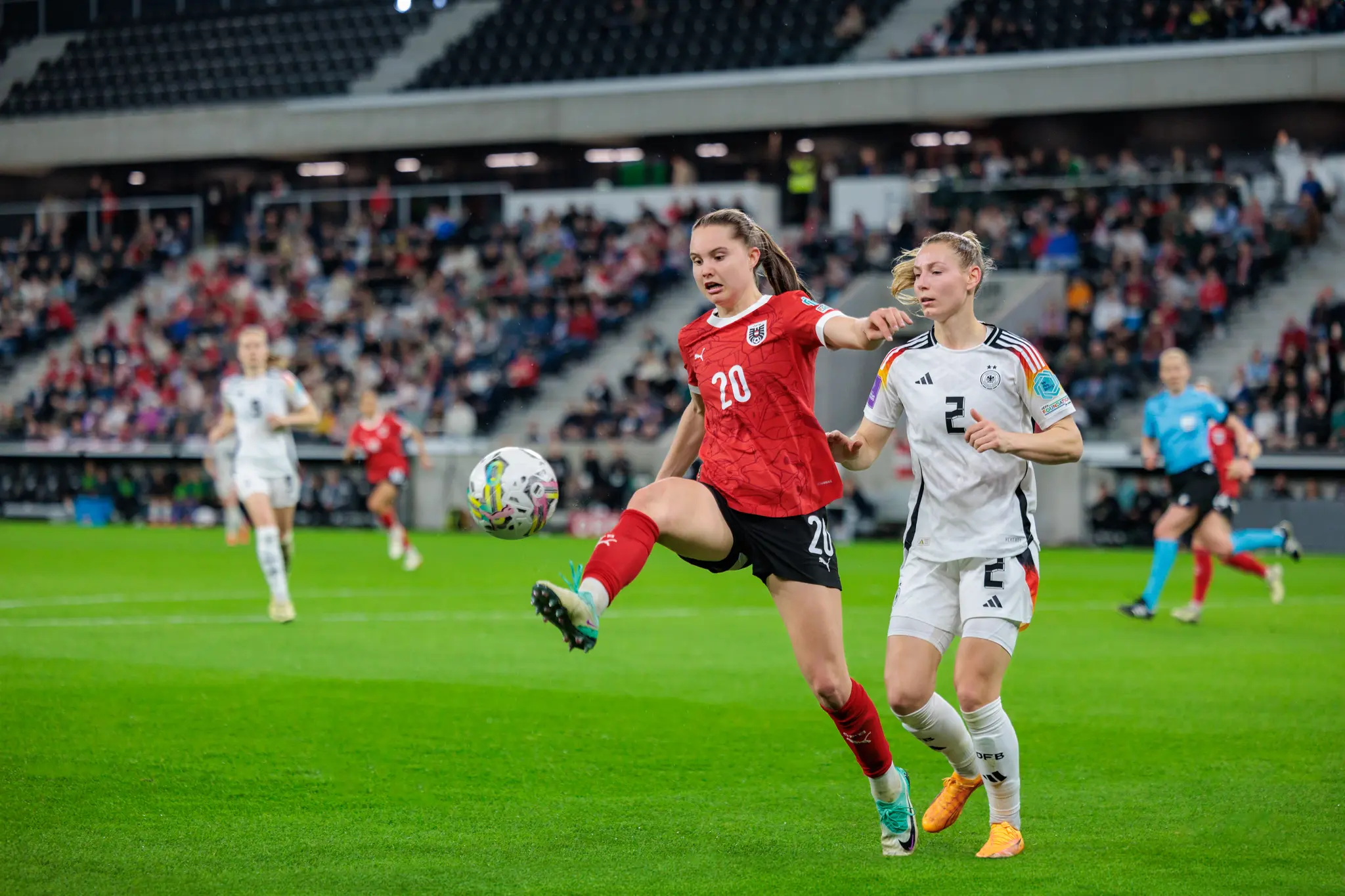 Eine österreichische Fußballspielerin im roten Trikot führt den Ball, verteidigt von einer deutschen Spielerin im weißen Trikot während eines Länderspiels vor Stadionzuschauern 