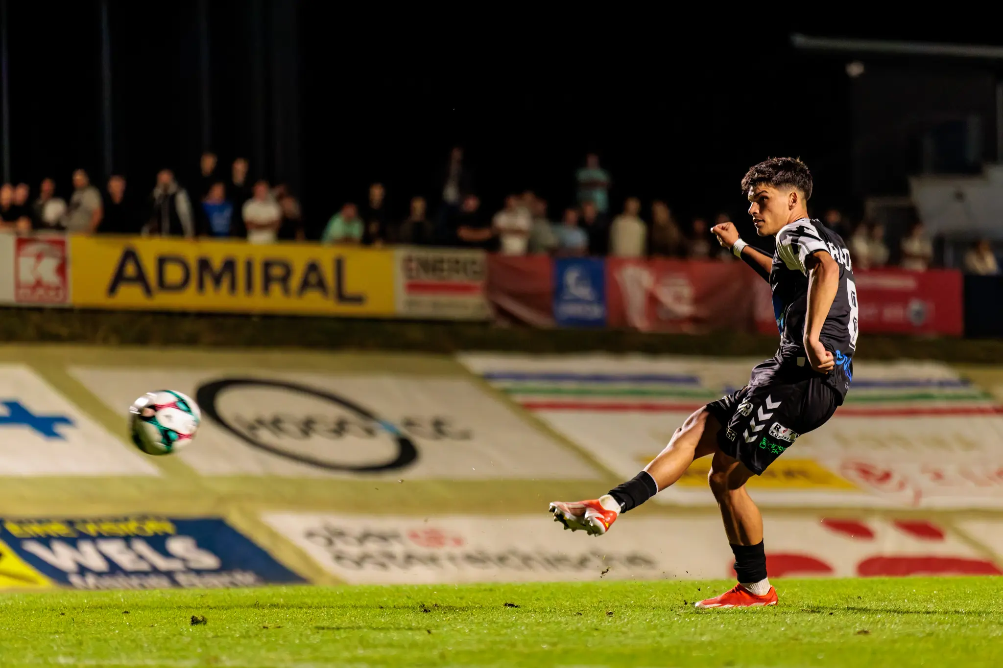 Ein Fußballspieler in schwarz-weißer Spielkleidung von SPG HOGO Wels tritt im ÖFB-Cup einen Elfmeter, wobei im Hintergrund eine Admiral-Werbebande zu sehen ist