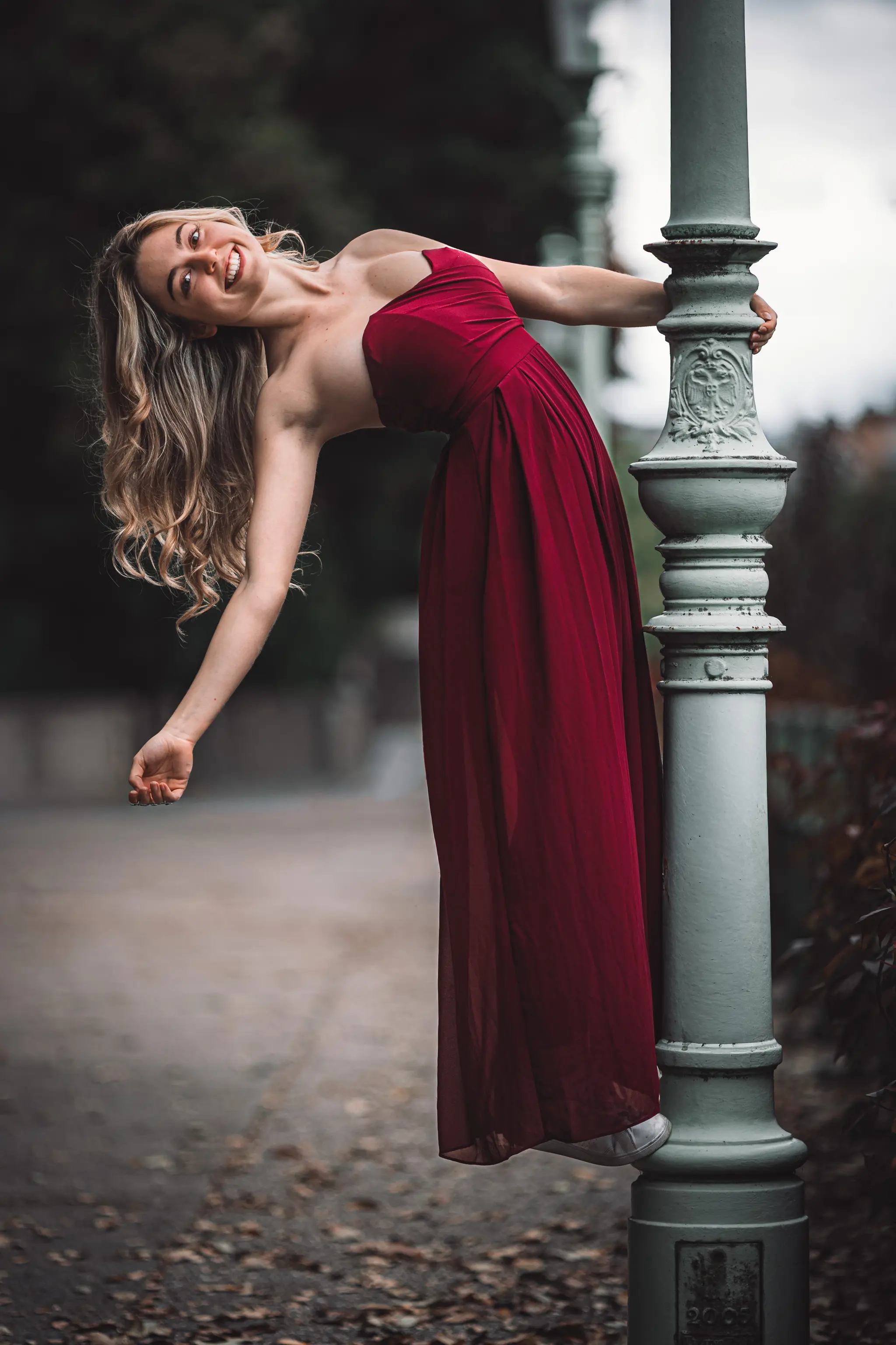 Model Valentina posiert bei einem Porträt-Shooting im Stadtpark Wien in einem eleganten roten Kleid und lehnt sich lachend an einen historischen Laternenmast zurück