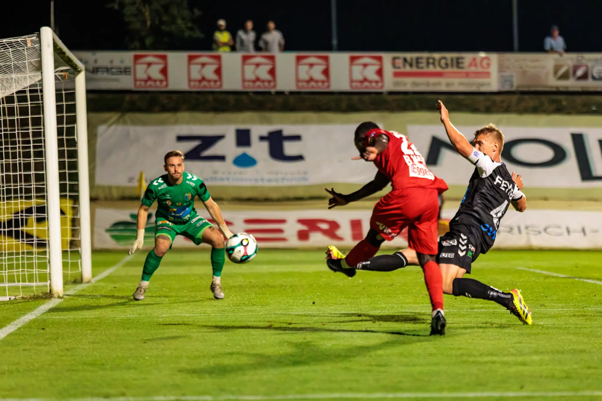 Ein Spieler des SKN St. Pölten in rotem Trikot schießt den Ball auf das Tor, während ein Welser Verteidiger in schwarz-weißem Trikot ihn zu blocken versucht und der Torwart zur Parade bereitsteht