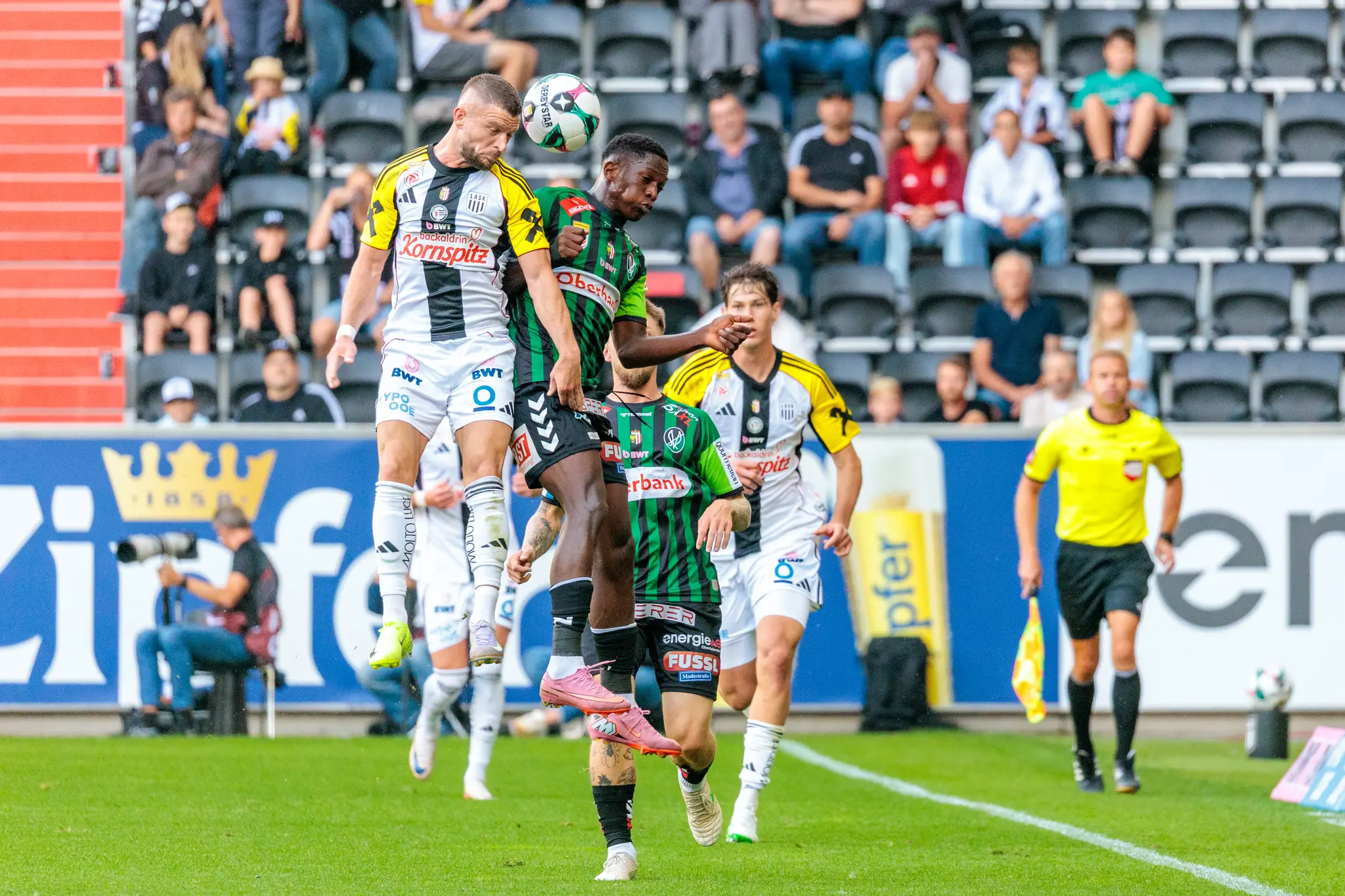 Ein Fußballspieler in einem schwarz-weiß-gelben LASK-Trikot und ein Spieler in einem grün-schwarz gestreiften Trikot der SV Ried springen gleichzeitig für einen Kopfball