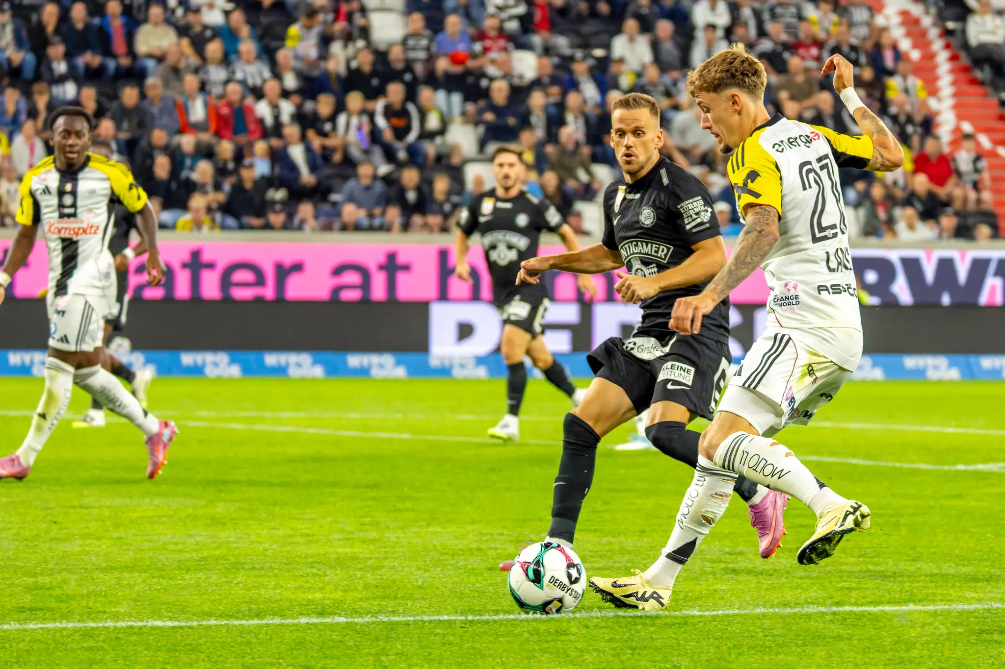 Zwei Fußballspieler, einer im hellen LASK-Trikot und einer im dunklen Trikot, ringen auf dem Spielfeld um den Ball vor einer Zuschauerkulisse