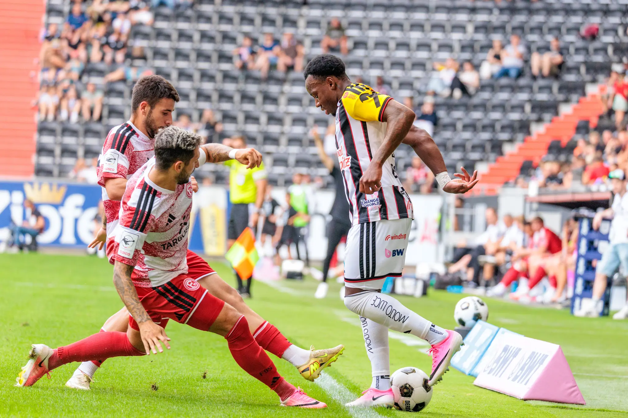 LASK-Spieler führt den Ball an der Seitenlinie gegen zwei Düsseldorfer Verteidiger, die Ersatzbank ist im Hintergrund zu sehen.