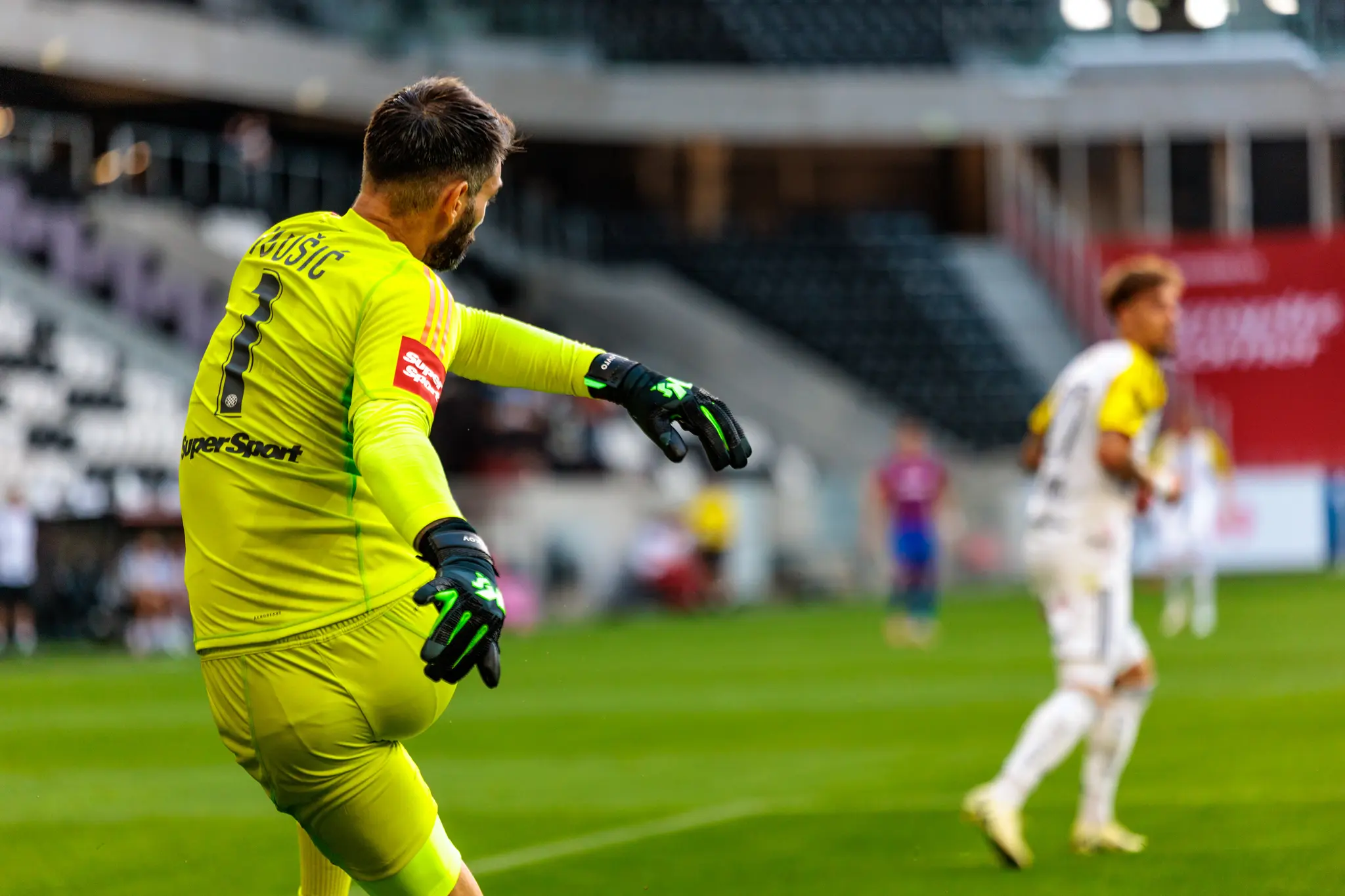 Ein Fußballtorhüter in leuchtend gelb-grünem Trikot mit der Nummer 1 und dem Namen Družić ist während eines Spiels in einem Fußballstadion zu sehen