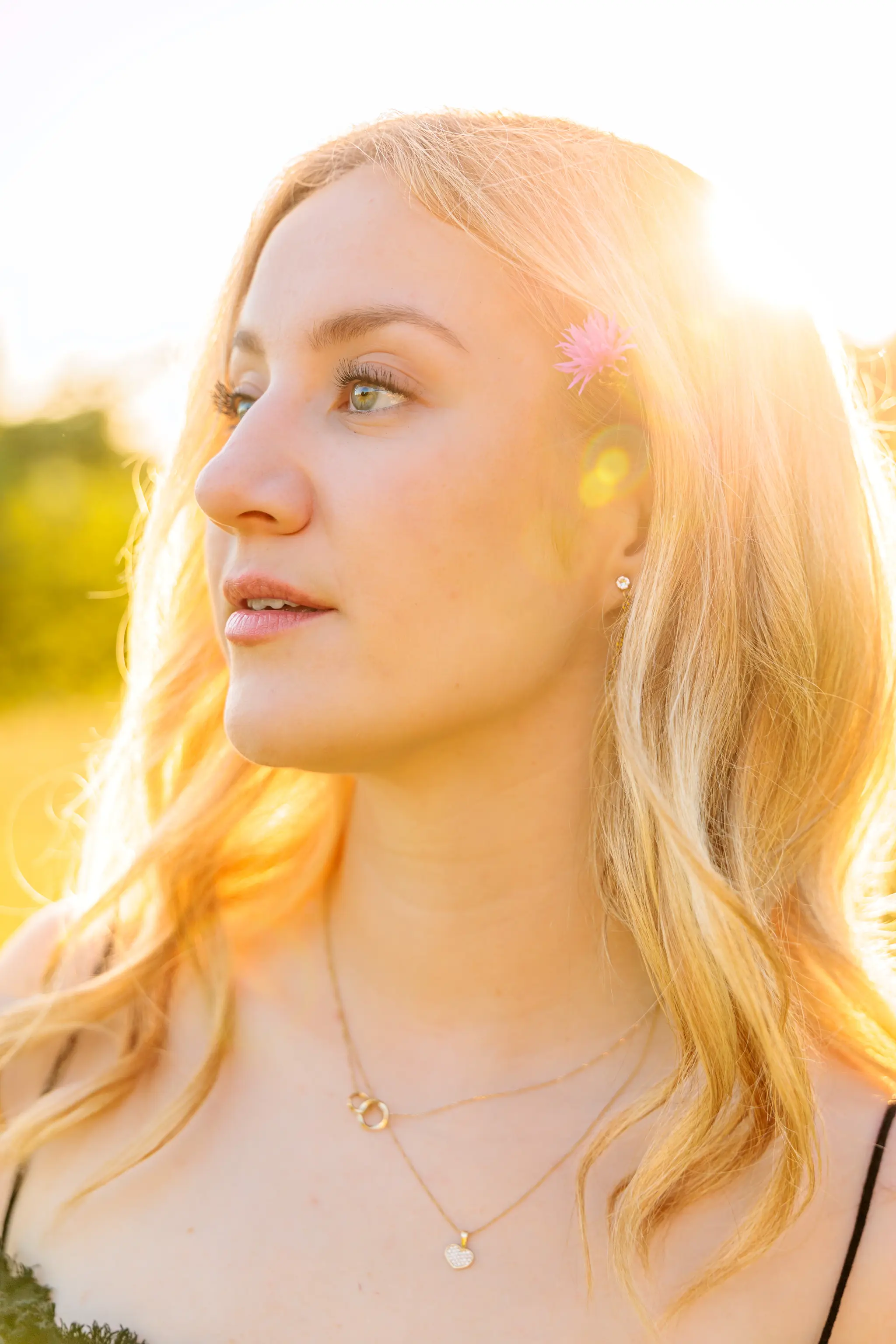 Nahaufnahme einer jungen blonden Frau mit rosa Blume im Haar, beleuchtet vom warmen goldenen Licht der Abendsonne in der Natur 
