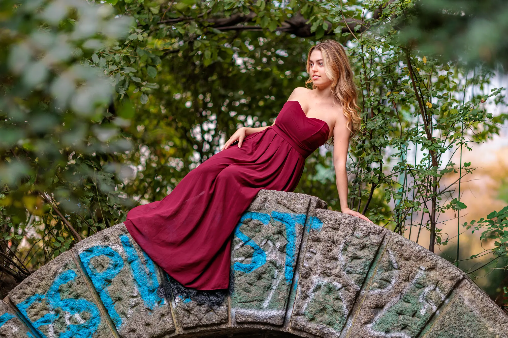 Model Valentina posiert während ihres Fotoshootings im Stadtpark elegant in einem roten Abendkleid auf dem Bogen einer alten Steinbrücke mit Graffiti