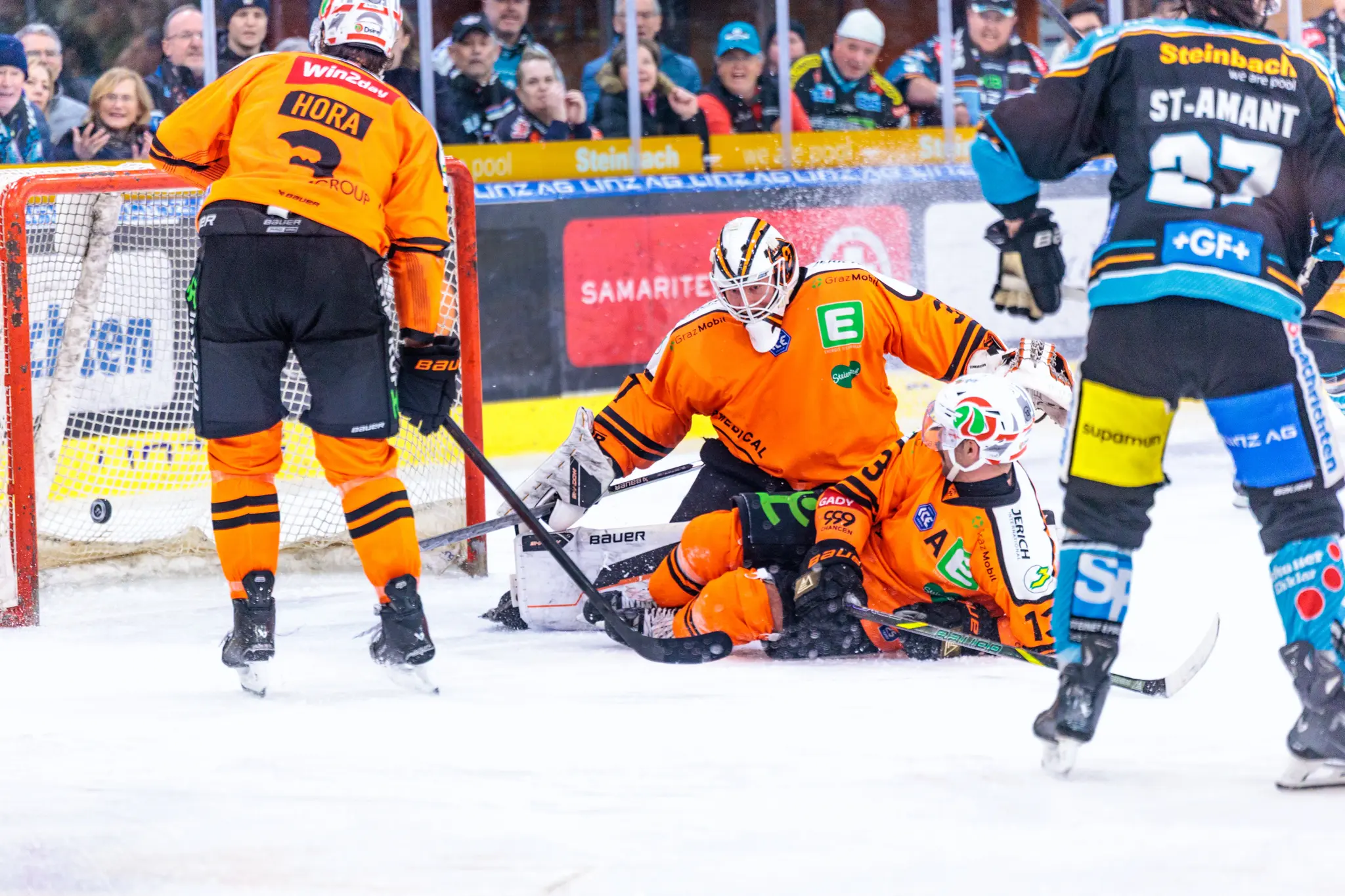 Eishockeyspieler in orangefarbenen und blau-schwarzen Trikots sind in einer dynamischen Spielsituation direkt vor dem Tor, wobei ein Spieler vor dem Torwart kniend am Eis liegt 