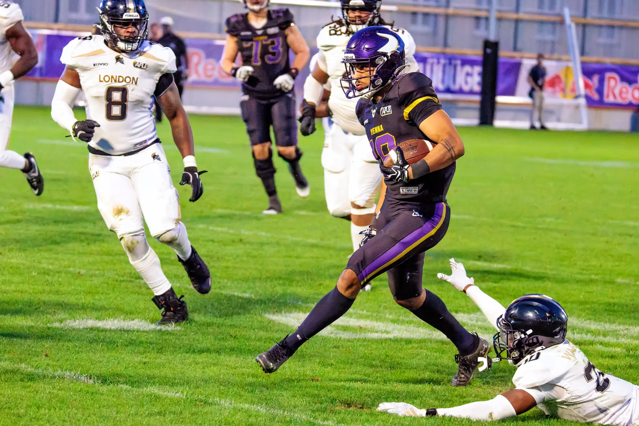 Ein Ballträger der Vienna Vikings bricht nach rechts durch, während ein London-Verteidiger hinter ihm zu Boden taucht – Laufspiel in der Schlussphase des dritten Viertels.