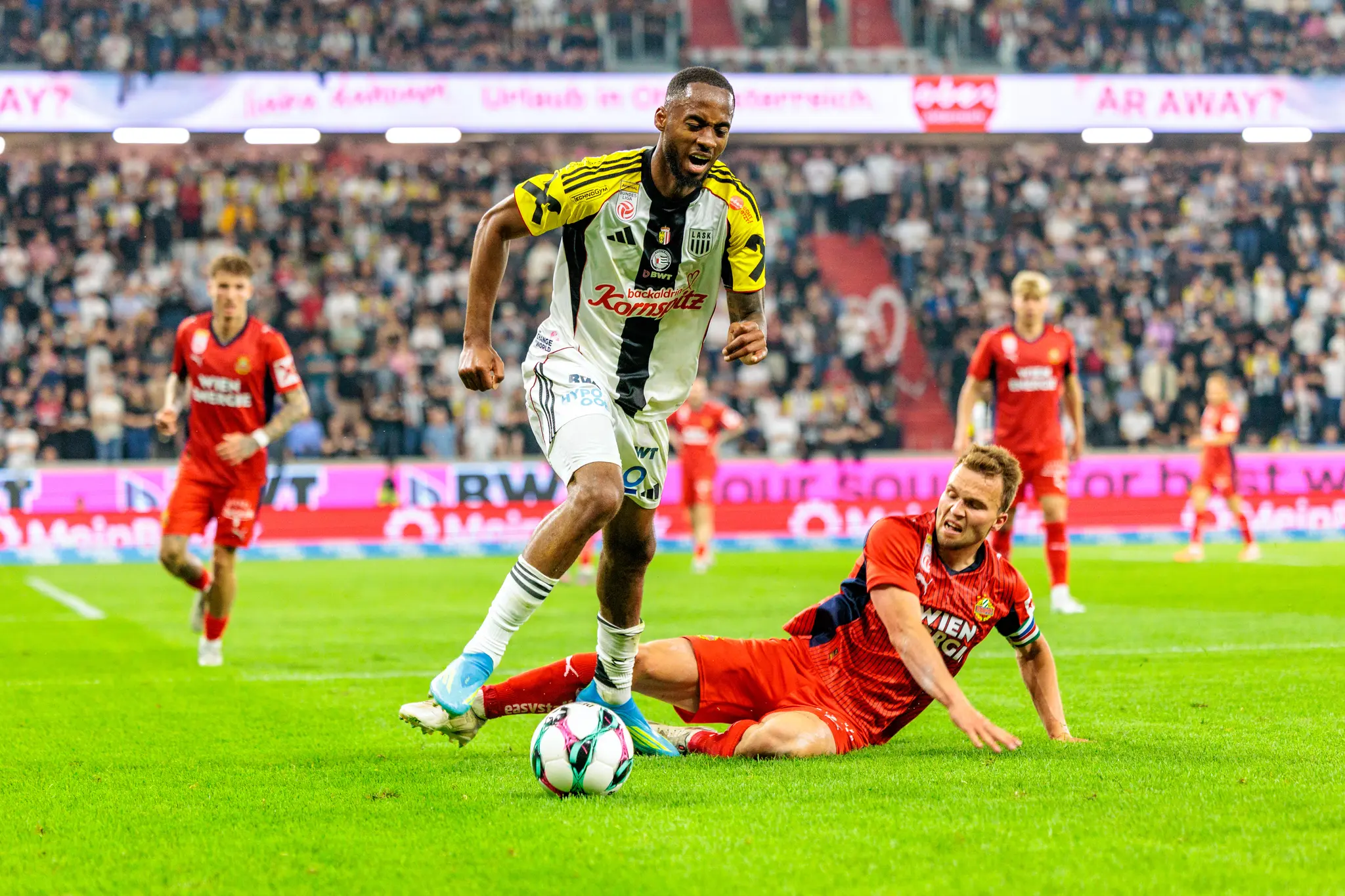 Ein Spieler des LASK führt den Ball an einem am Boden grätschenden Spieler des SK Rapid vorbei