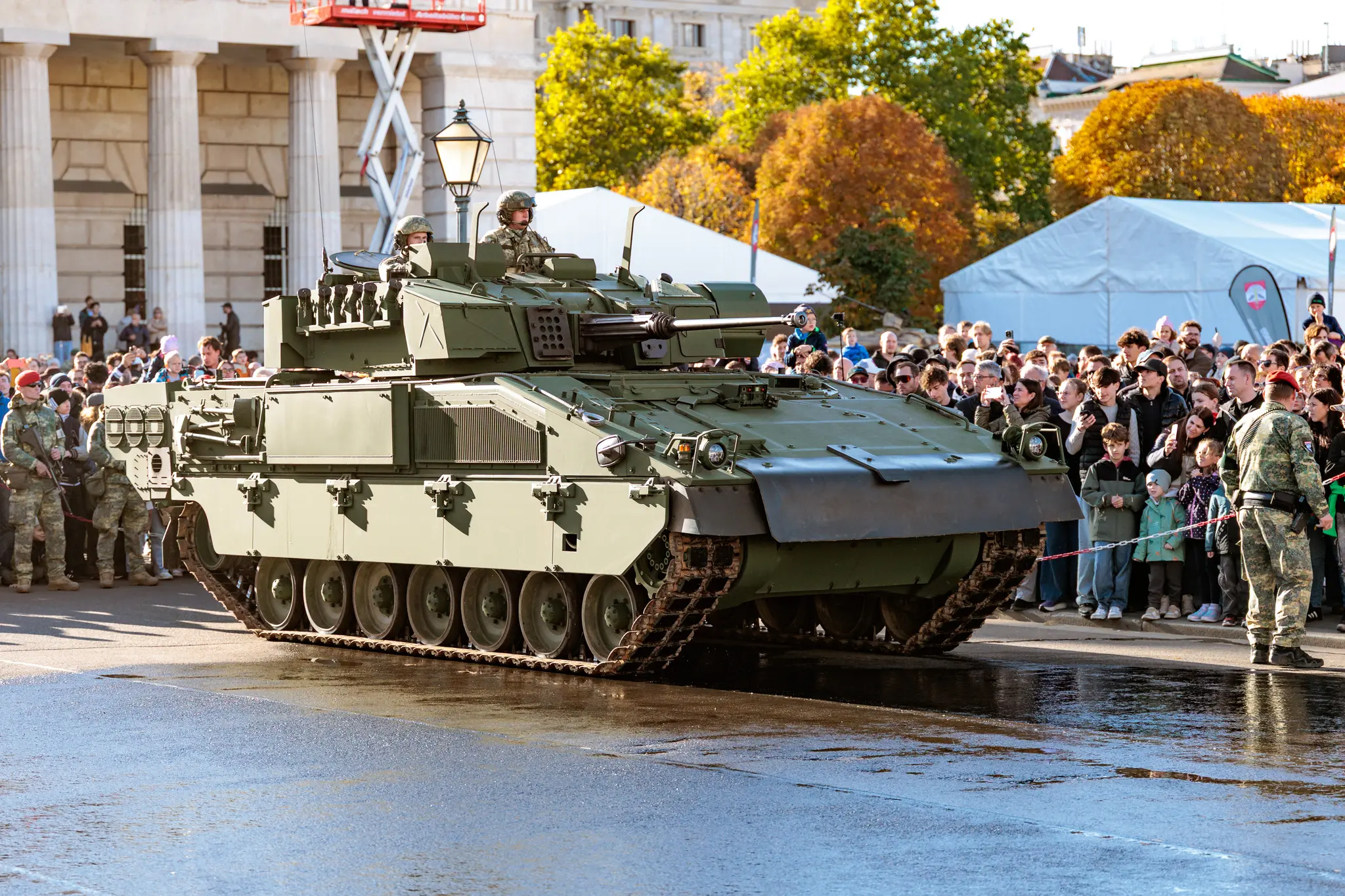 Ein Schützenpanzer Ulan des österreichischen Bundesheeres rollt über den nassen Asphalt des Heldenplatzes, hinter einer Absperrung verfolgt das Publikum die Fahrt.