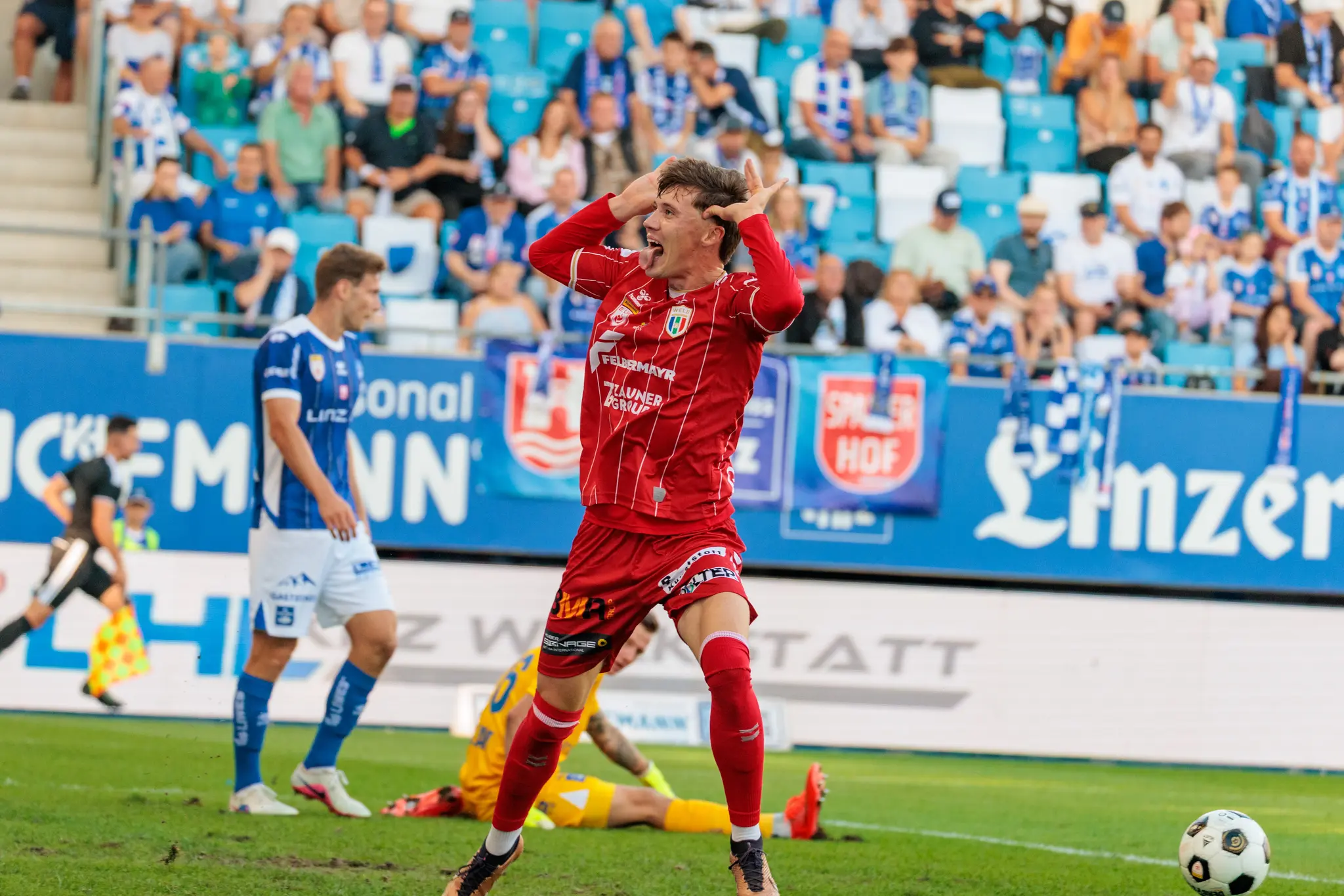 Der Welser Torschütze Sebastian Malinowski feiert ausgelassen vor der blau-weiß besetzten Heimtribüne: beide Hände an den Schläfen, Mund und Zunge herausgestreckt; im Hintergrund die Linzer Fans.