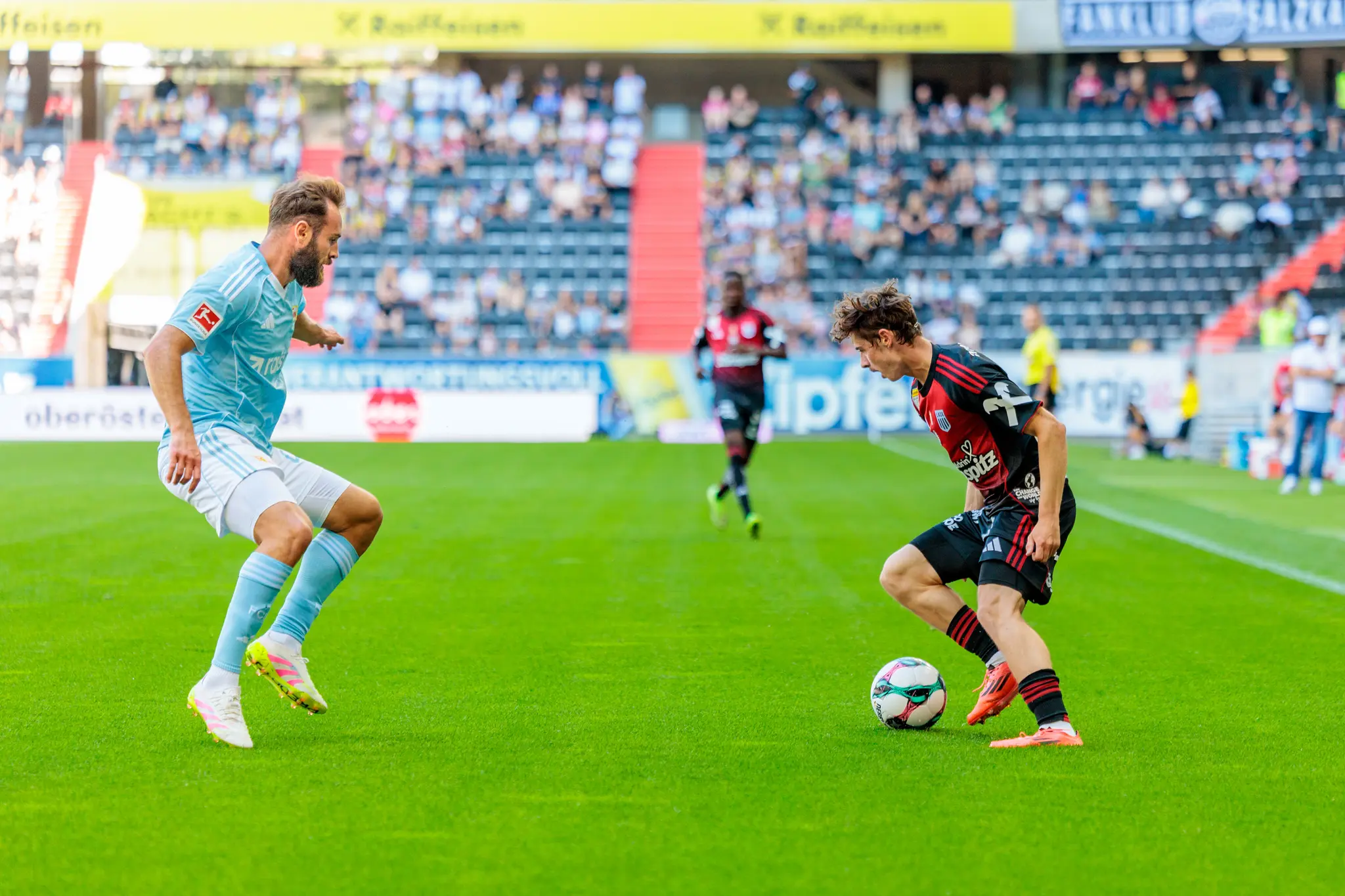 Ein Fußballspieler in hellem Trikot blickt auf seinen Gegner in dunklerem Trikot, der den Ball am Fuß hat, während eines Spiels vor Stadionpublikum