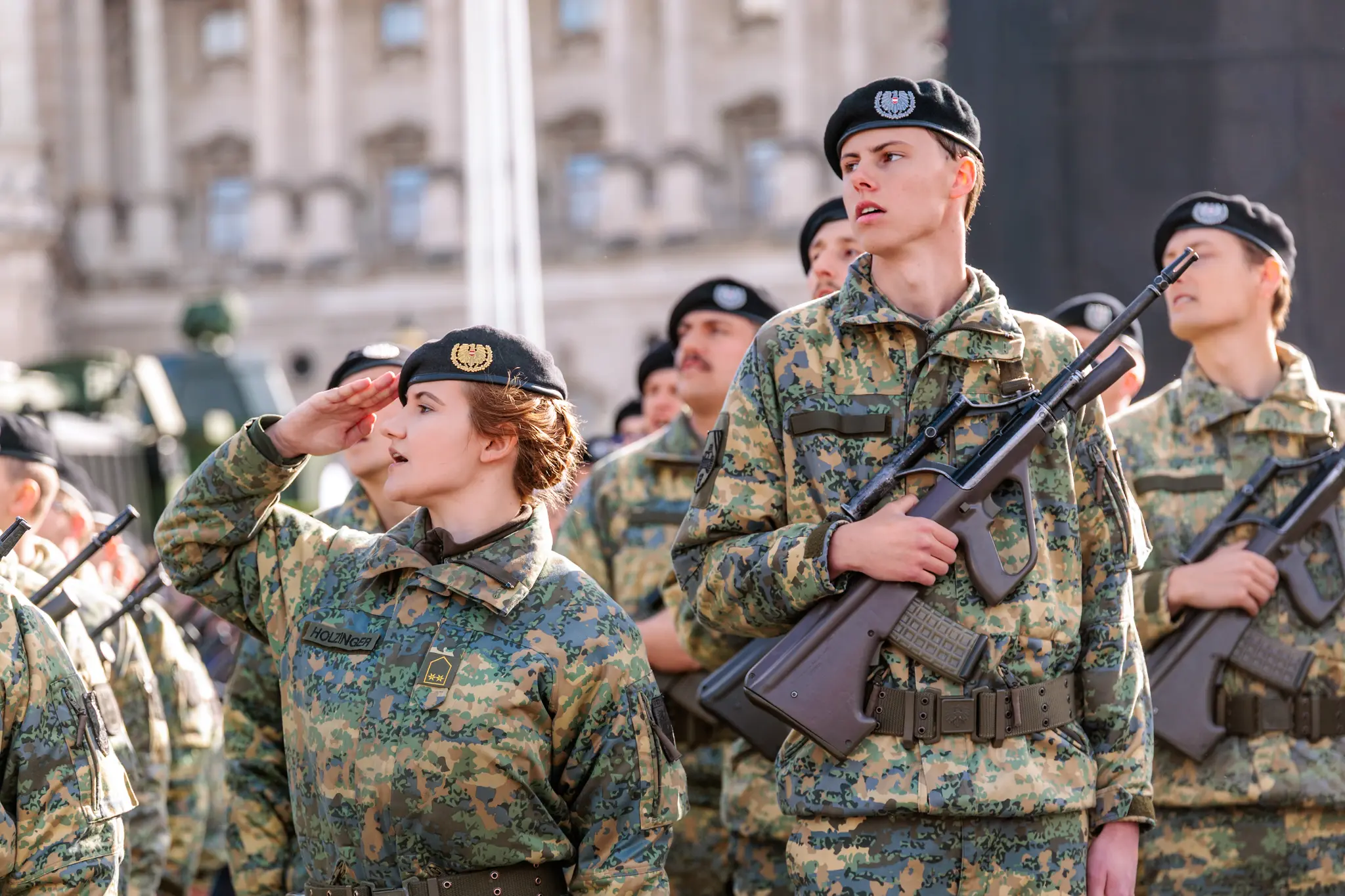 Eine Kommandantin in Tarnuniform salutiert mit offenen Augen, neben ihr steht ein Rekrut mit Sturmgewehr.