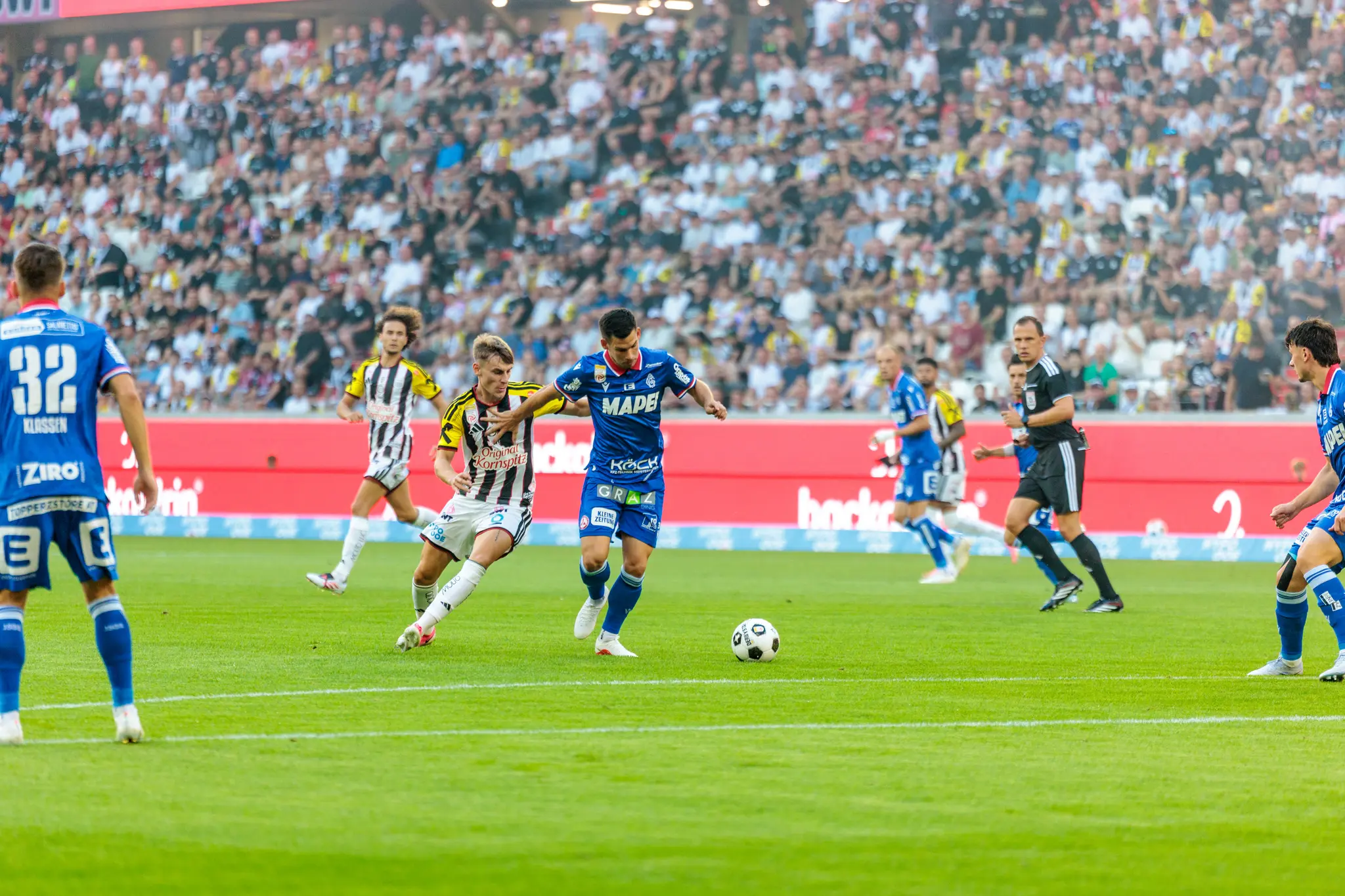 Zweikampf zwischen einem LASK-Spieler im schwarz-weiß-gelben Trikot und einem GAK-Verteidiger im blauen Trikot um den Ball in der Nähe der Spielfeldmitte