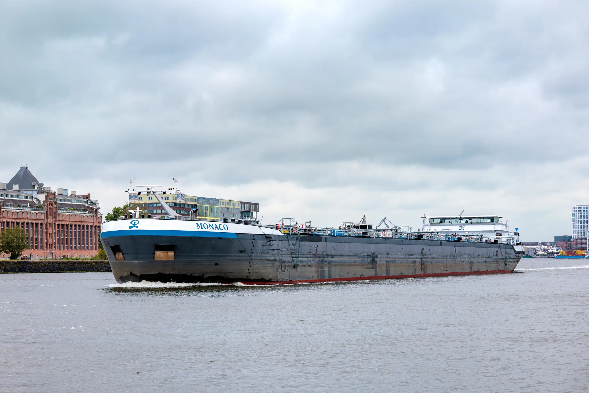 Ein großes, graues Frachtschiff namens Monaco befährt unter einem bewölkten Himmel einen Wasserweg in Amsterdam, gesäumt von verschiedenen Stadtgebäuden