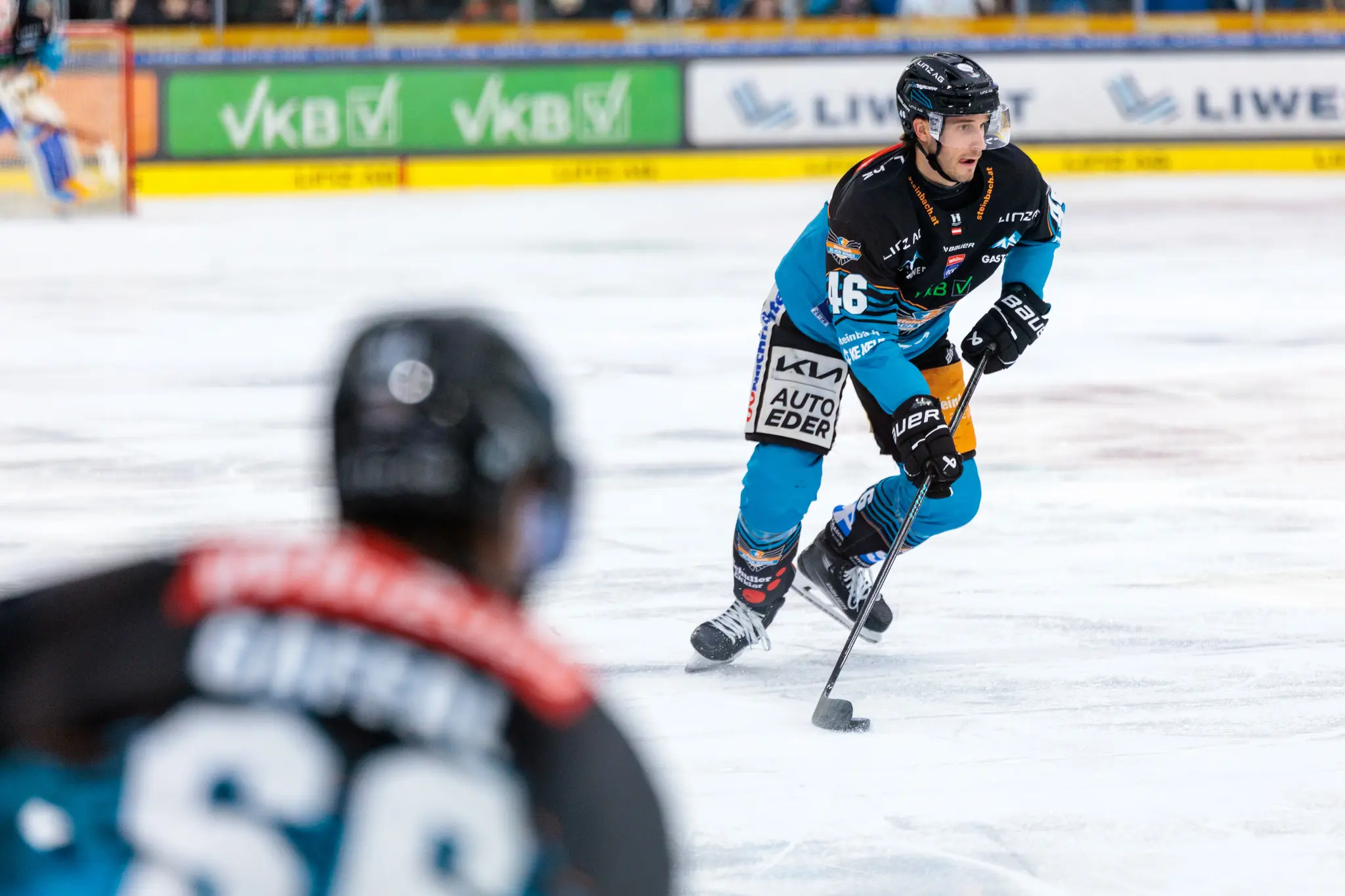 Ein Eishockeyspieler der Black Wings Linz in schwarzer und türkiser Uniform fährt auf dem Eis und hat den Puck am Schläger