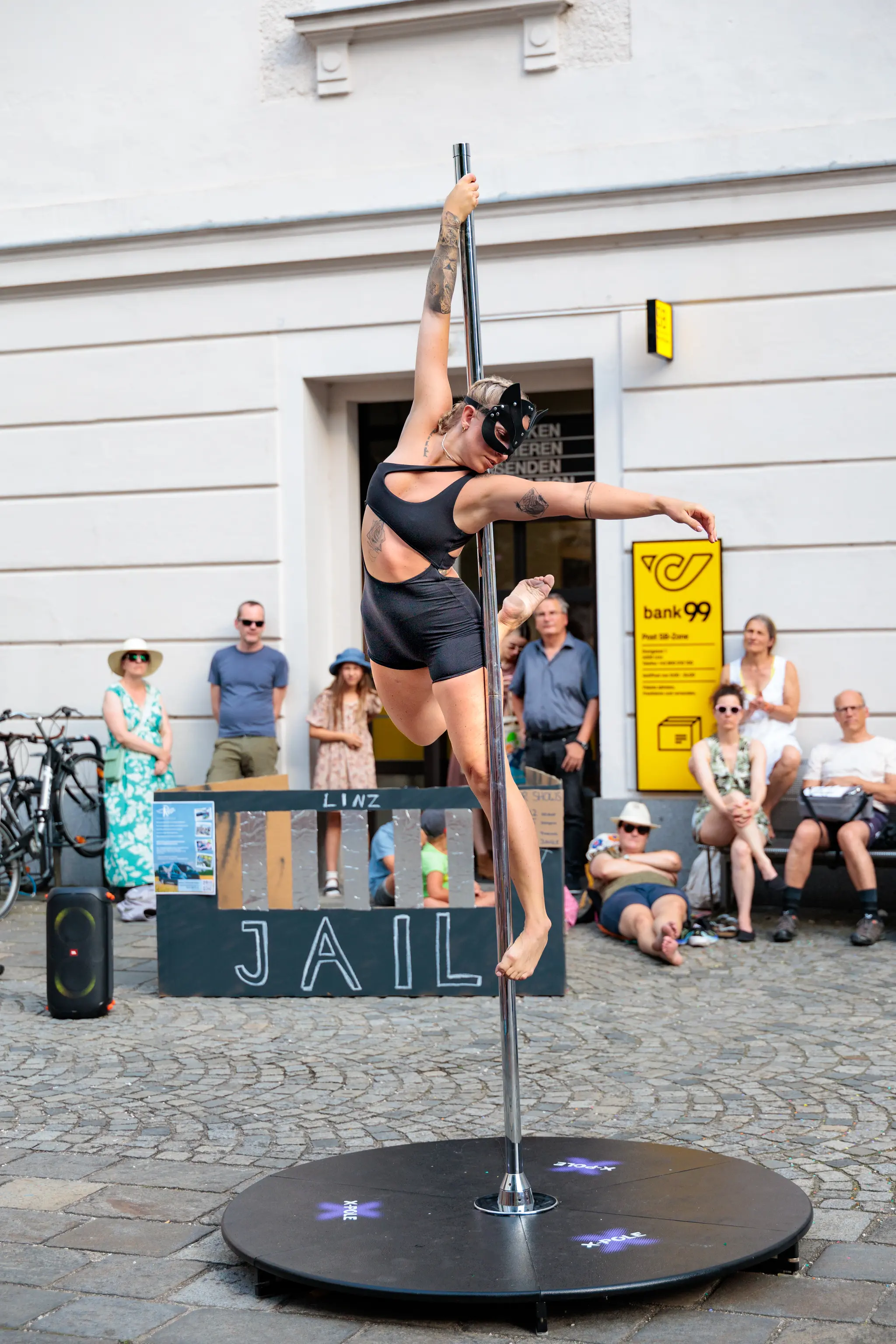 Eine Frau in schwarzem Poledance-Outfit und Katzenmaske zeigt eine akrobatische Pose an einer Outdoor-Stange. Im Hintergrund beobachten Zuschauer die Performance beim Pflasterspektakel 2024 in Linz 