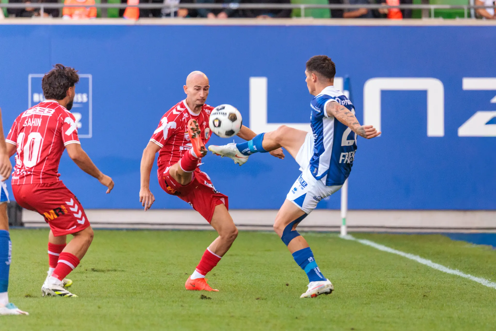Dominik Frieser vom FC Blau-Weiß Linz und ein Welser Spieler liefern sich an der Seitenlinie ein hohes Bein-Duell, der Ball springt zwischen ihnen auf; im Hintergrund leuchtet die Linz-AG-Bande.