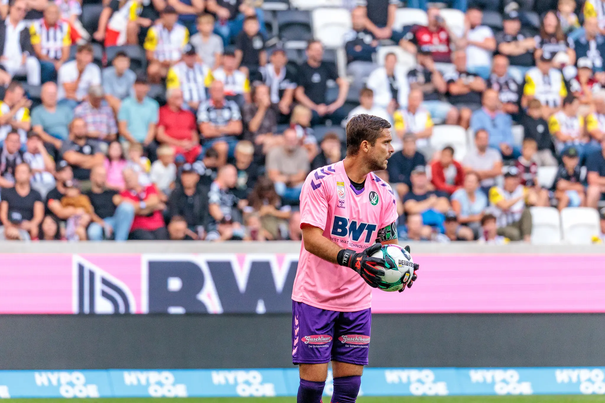 Amdreas Leitner im pinken BWT hält den Spielball während eines Derbyspiels gegen den LASK, mit unscharfen Fans und einer BWT Bande im Hintergrund