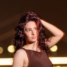 Eine junge Frau mit voluminösem, lockigem und rötlich-braunem Haar fährt sich mit der Hand durch die Haare und schaut nachdenklich nach oben